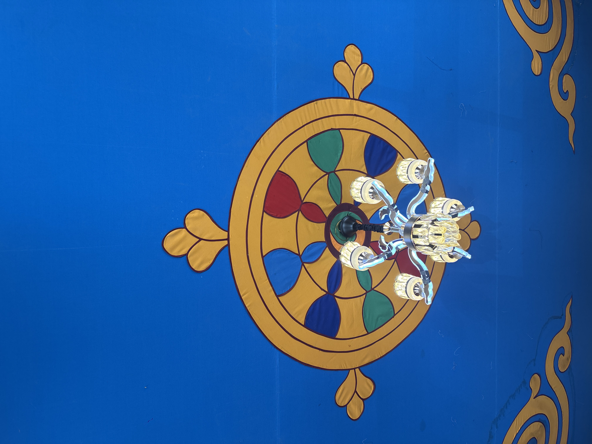 The image shows a low-angle shot of a ceiling, likely in a building in Jila, China. The ceiling is painted a vibrant blue. A large, decorative Dharma wheel is centered in the frame, featuring a yellow outer ring with stylized leaf-like protrusions extending outwards at four points. The interior of the wheel is divided into segments filled with red, green, and blue colors. A modern chandelier with multiple lights is suspended from the center of the Dharma wheel, partially obscuring the wheel's central details. A gold decorative element is visible in the top-right corner of the frame.