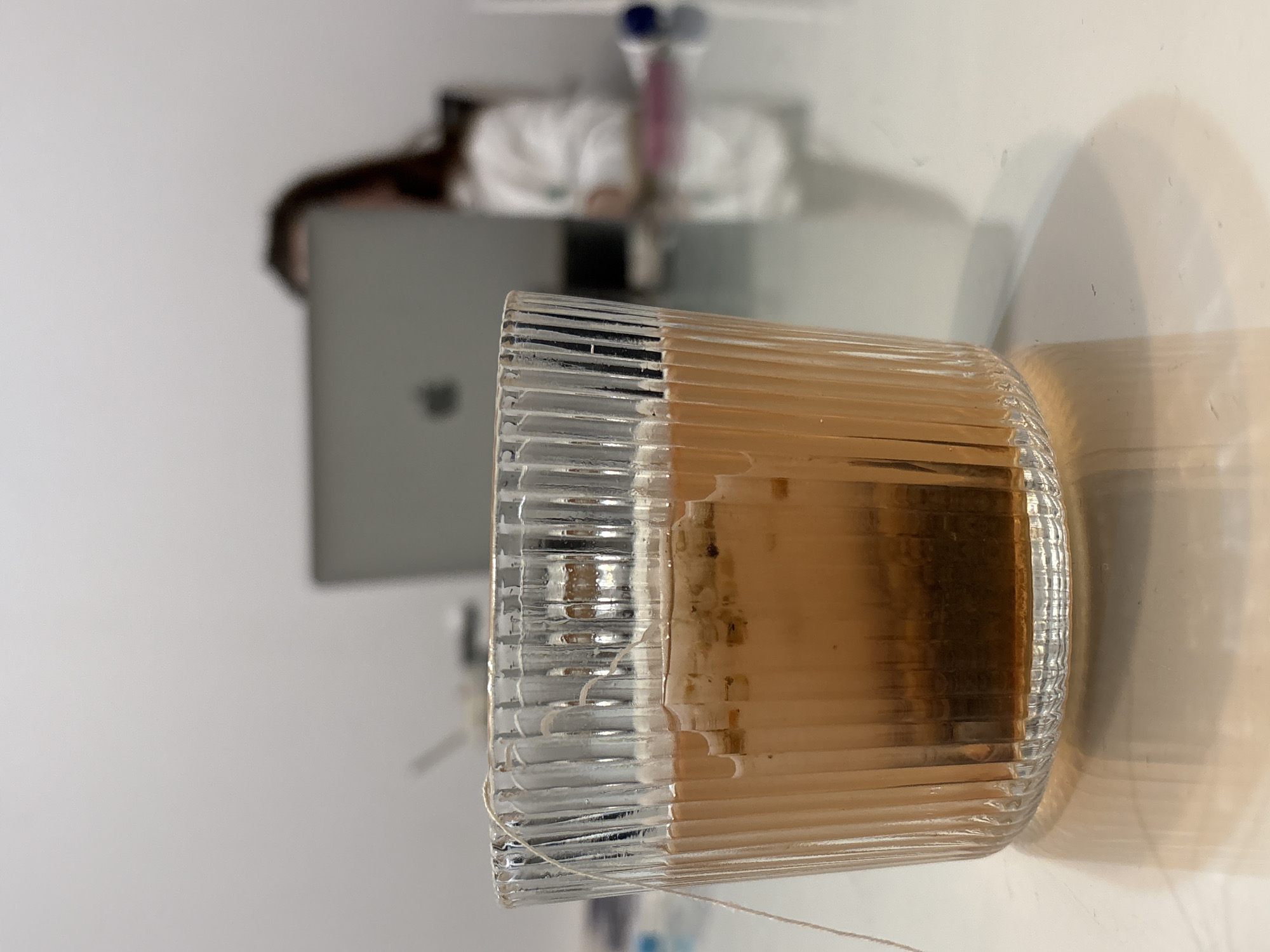 A lit candle in a ridged glass container sits on a white surface. The candle appears to be mostly consumed. In the background, out of focus, is a laptop and a person, possibly working. The location is likely indoors but there is no definitive evidence to identify the location as Kragujevac, Serbia.