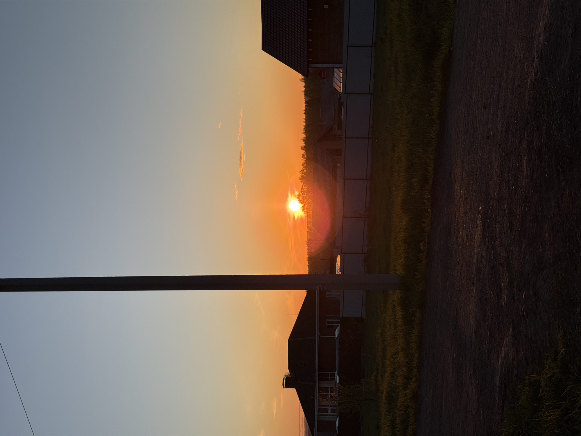 A sunset over Старые Кены, Russia, illuminates the horizon, casting a warm orange glow across the sky. Silhouetted against the setting sun are residential buildings and a telephone pole. A road borders one side of the frame.