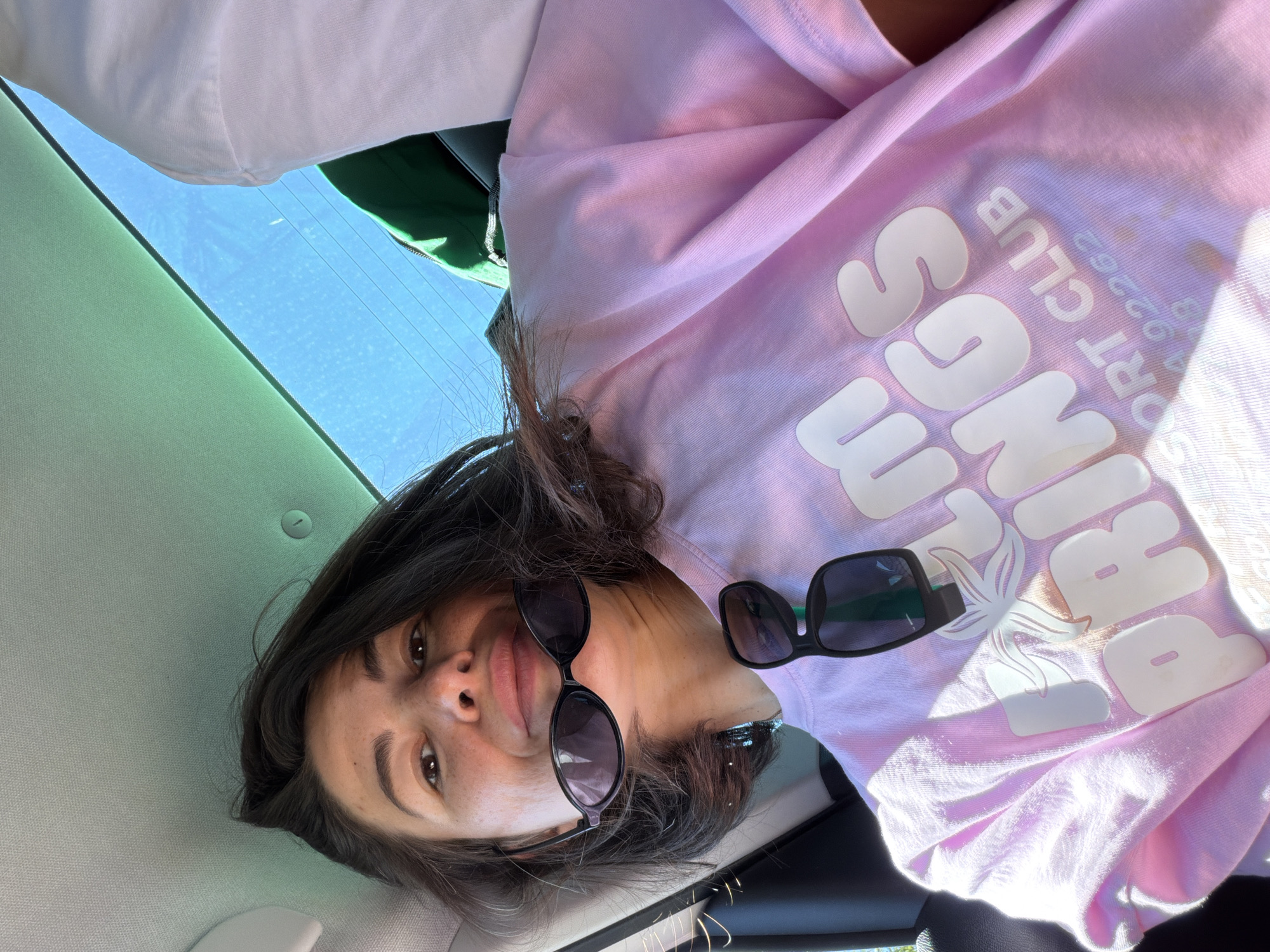 The image captures a casual selfie of a person inside a car, taken on a bright, clear day in Kubasi, Montenegro. The photograph is oriented on its side, with the person's head to the left and their body extending to the right of the frame.
The central subject is a person with dark hair, looking directly at the camera with a neutral, slightly pouting expression. A pair of black sunglasses is worn pushed up on their face, and a second pair of black sunglasses hangs from the neckline of their shirt. The person is wearing a light pink, short-sleeved t-shirt.
Notable details on the t-shirt include large, white, rounded block letters spelling "SPRINGS". Above this, smaller white text reads "SPORT CLUB" followed by the number "02262". A white graphic, resembling a stylized starfish or a tropical leaf, is also visible on the shirt.
The setting appears to be a car interior. Above the person's head (which would be the ceiling if the image were rotated correctly), a light-colored car headliner is visible. To the right side of the frame, a clear, bright blue sky is visible through a window, indicating sunny weather during daytime. A glimpse of a dark green object, possibly a bag or another piece of clothing, can be seen behind the person's right shoulder. The activity is a self-portrait taken in a relaxed moment within the vehicle.