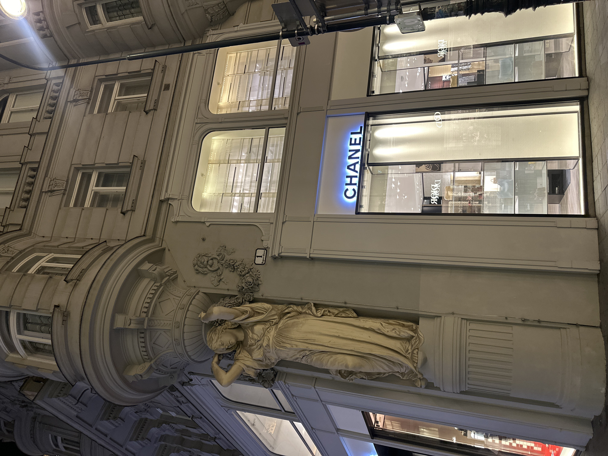 The image shows the exterior of the Chanel boutique in Vienna, Austria, located at Kohlmarkt 14. The Chanel sign, illuminated with blue neon lights, is prominently displayed above the entrance. The boutique features large glass windows that showcase the interior display areas. To the left of the Chanel entrance, a classical statue of a woman holding a wreath adorns the facade of the building. This statue is positioned on a decorative pillar, and intricate architectural details, including ornate carvings and window frames, are visible on the building's exterior. The street and pavement are visible in the foreground, and a lamppost with a bright light is positioned to the right of the building. The scene is captured at night, with artificial lighting illuminating the building and its surroundings.