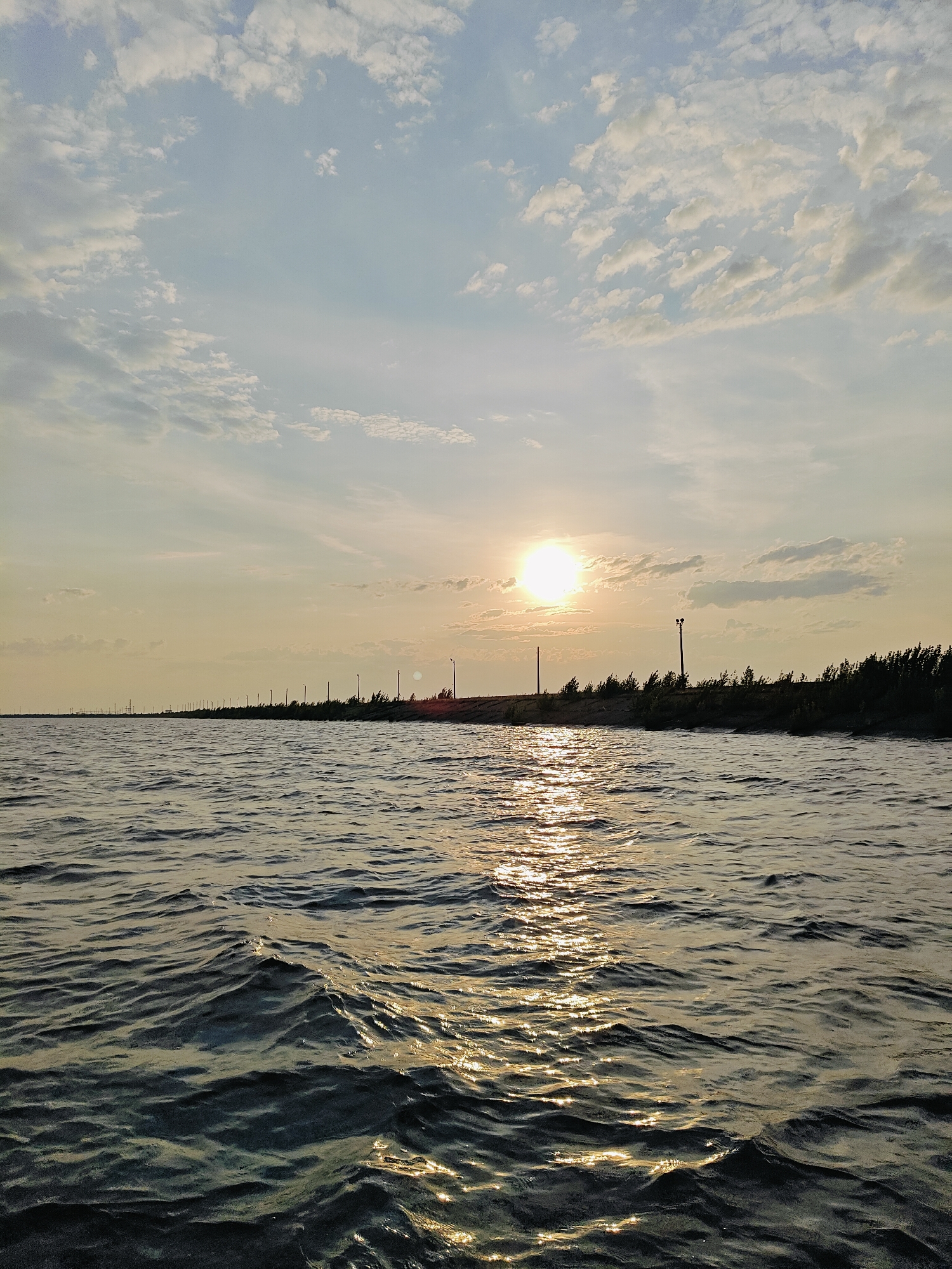 A serene landscape captures the golden hour over a calm body of water, likely the Kama River in Chaikovsky, Russia. The sun, a bright orb, hangs low in the hazy sky, casting a warm, diffused light and creating shimmering reflections on the water's surface. Scattered, wispy clouds drift across the pale blue expanse. Along the distant horizon, a line of silhouetted trees marks the edge of the land, punctuated by a series of tall, slender utility poles that stretch into the distance. The water in the foreground exhibits gentle ripples, catching the sunlight in sparkling patterns, suggesting a slight breeze. The overall impression is one of tranquility and natural beauty at the close of day.