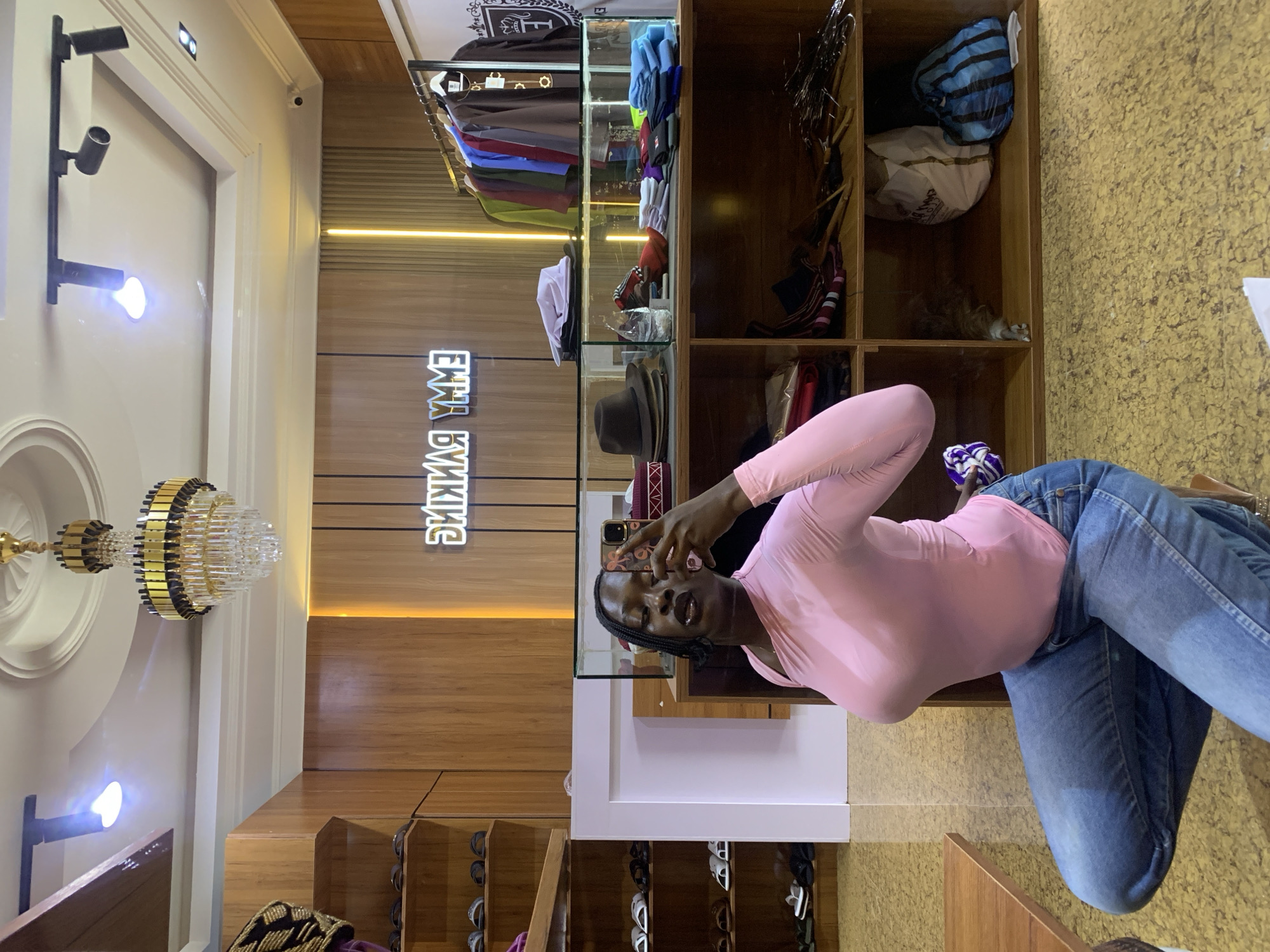 A young Black woman is taking a mirror selfie in a clothing store. She is wearing a pink long-sleeved top and blue jeans. She is kneeling and holding her phone up to take the photo. The store has wooden shelves filled with clothing, hats, and bags. There is a neon sign on the wall that reads "EMMY BANKING". The store appears to be located in Gura Top, Nigeria.