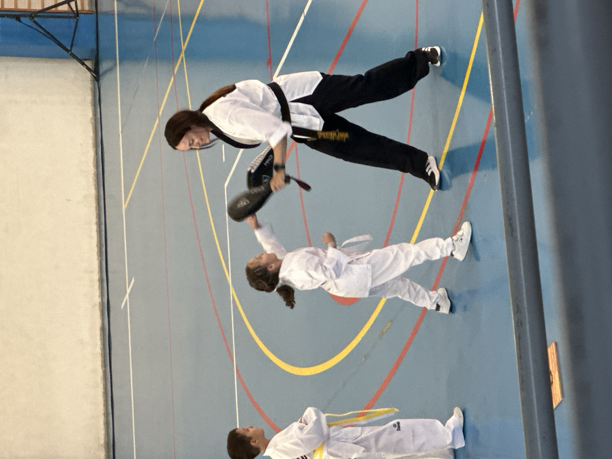 Here is a description of the image:
A slightly skewed, full indoor shot depicts three individuals engaged in martial arts practice on a blue-floored gymnasium. The floor is marked with yellow, white, and red lines.
One adult female, presumably an instructor, is dressed in a white martial arts uniform with a black belt and black pants. She is holding a black target in her left hand, positioned to be struck.
A young child, wearing a white martial arts uniform with a white belt, is performing a striking kick at the target held by the adult female.
Another young child is lying on the floor, wearing a white martial arts uniform with a yellow belt. The child is facing upwards.
The location is unknown, as there are no discernible details to identify the city or specific location. The setting is clearly an indoor gymnasium used for martial arts practice.