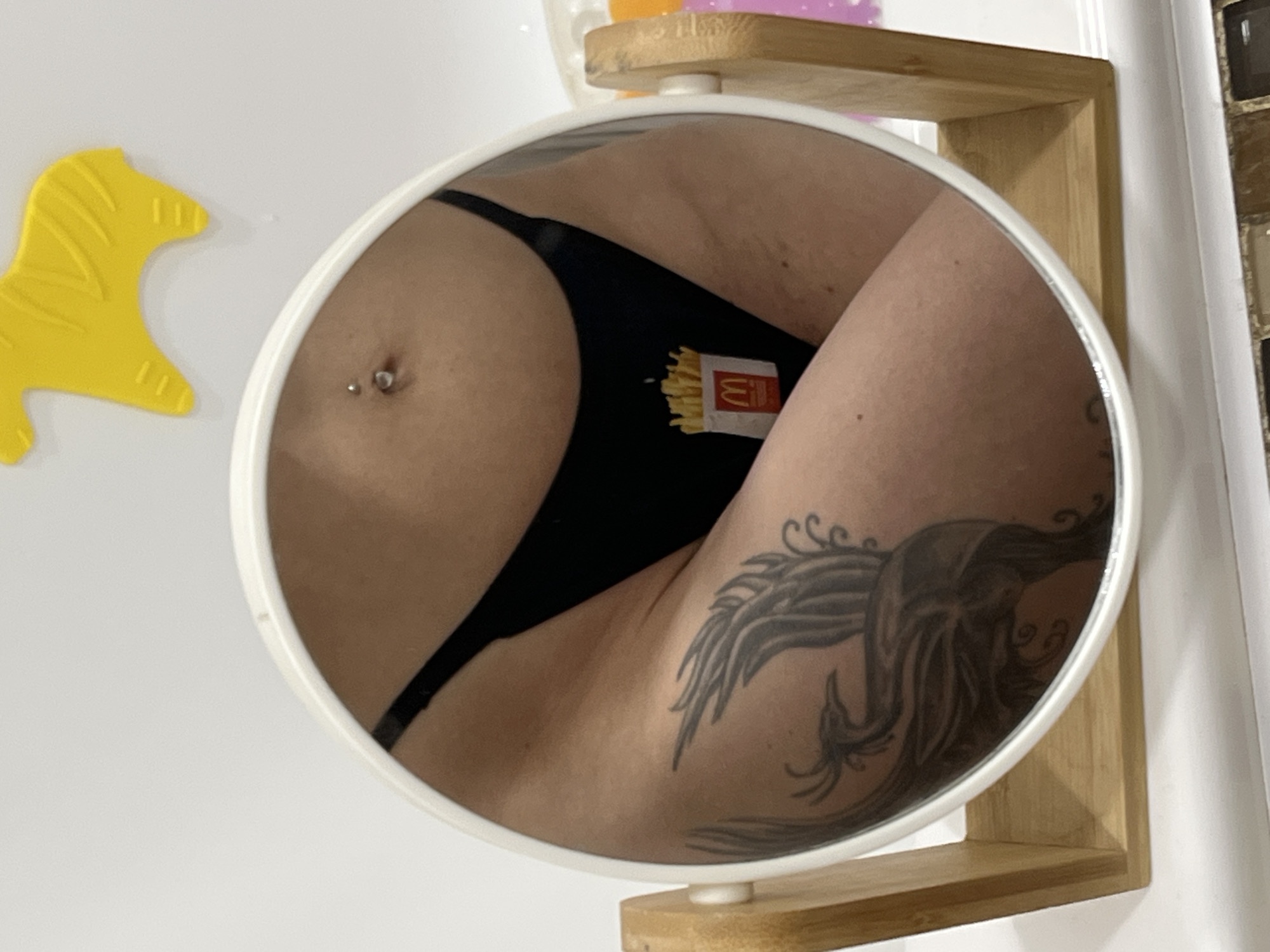 The image is a close-up, reflective shot of a person's lower torso and abdomen, captured in a white-rimmed, oval mirror with a wooden stand. The mirror is positioned on a white surface that appears to be a countertop, possibly in a bathroom. A yellow, stylized horse-shaped decoration is visible in the upper right background, out of focus.
The subject is wearing black underwear with a print of McDonald's french fries. A navel piercing is visible on the subject's abdomen. A large tattoo of a phoenix in shades of grey covers the subject's thigh and hip. The lighting is bright, casting soft shadows.
There is no indication of the image being located in Nizhny Novgorod, Russia.
