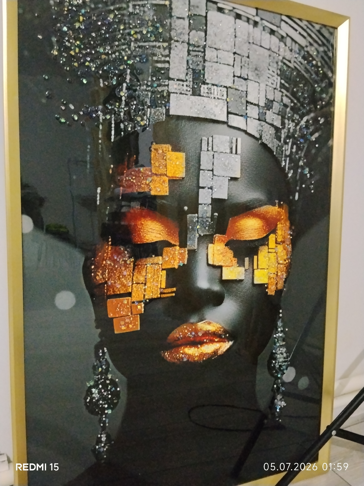 An art piece featuring a woman's face is depicted. The artwork is framed in gold and hangs on a white wall. The woman's face is a dark color, and her eyes are adorned with mosaic-like squares of orange and gold. Her lips are also decorated with a similar mosaic pattern in shades of gold. Her hair is a mix of dark strands with glitter and metallic silver squares. She wears large, dangly earrings that are also made of dark, sparkling elements. The background of the artwork is dark and appears to have a subtle gradient. There are reflections on the surface of the artwork. A black metal stand is partially visible in the lower right corner. The bottom left corner of the image shows "REDMI 15" in white text, and the bottom right corner displays a date and time stamp: "05.07.2026 01:59". The image is a photograph taken from a slight angle, showing the texture and shimmer of the artwork.