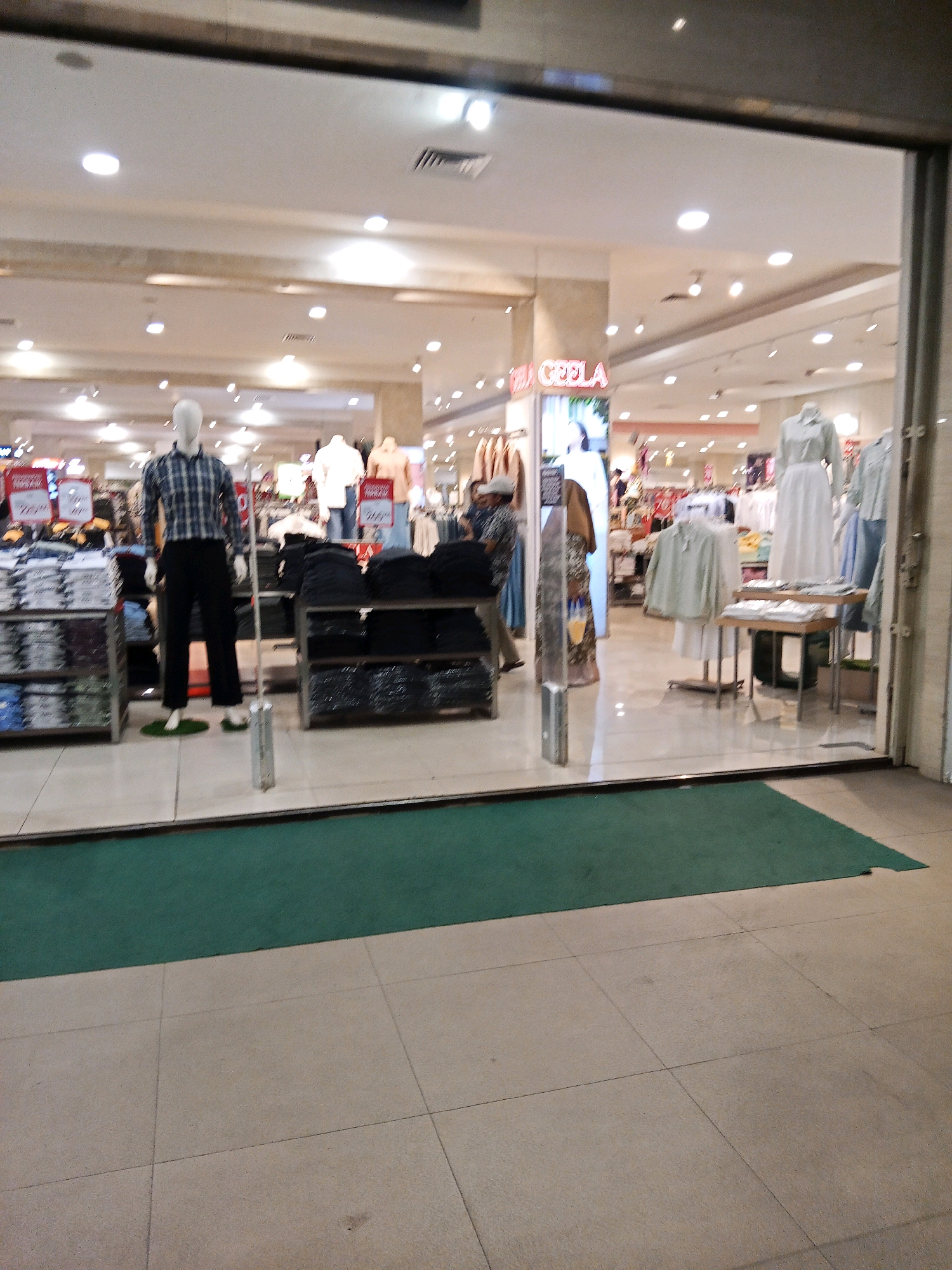 The image shows the entrance to a clothing store, identified as "CELANA" by signage, located in a mall in Lubuklinggau, Indonesia. The store's interior is visible through a large glass storefront, revealing displays of clothing on shelves and mannequins. In the foreground, a green carpet leads into the store, with a tiled floor beyond. A male mannequin stands in the center, dressed in a checkered shirt and black pants, flanked by racks of folded clothing. To the right, more clothing displays are visible, including dresses and shirts on mannequins. The store appears well-lit with recessed ceiling lights.