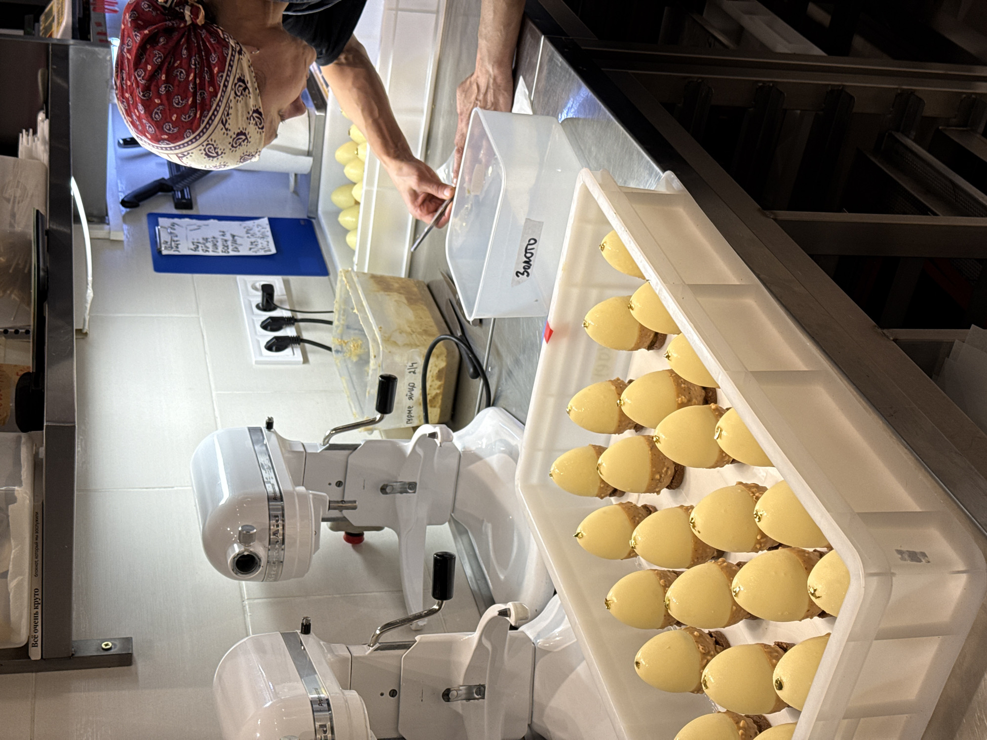 The image shows an indoor kitchen environment, possibly a professional bakery or pastry shop, in Belgrade, Serbia. A person wearing a red and white patterned headscarf is focused on a task involving a container of lemon-shaped pastries with gold leaf accents. Another container labelled "Золото" (Gold in Russian) is present. The scene includes food preparation equipment such as stand mixers. The pastries appear to be arranged in a white plastic tray. There is a clipboard with handwritten notes and a power outlet with plugs. The environment appears clean and organized. Labels written in Russian are evident.