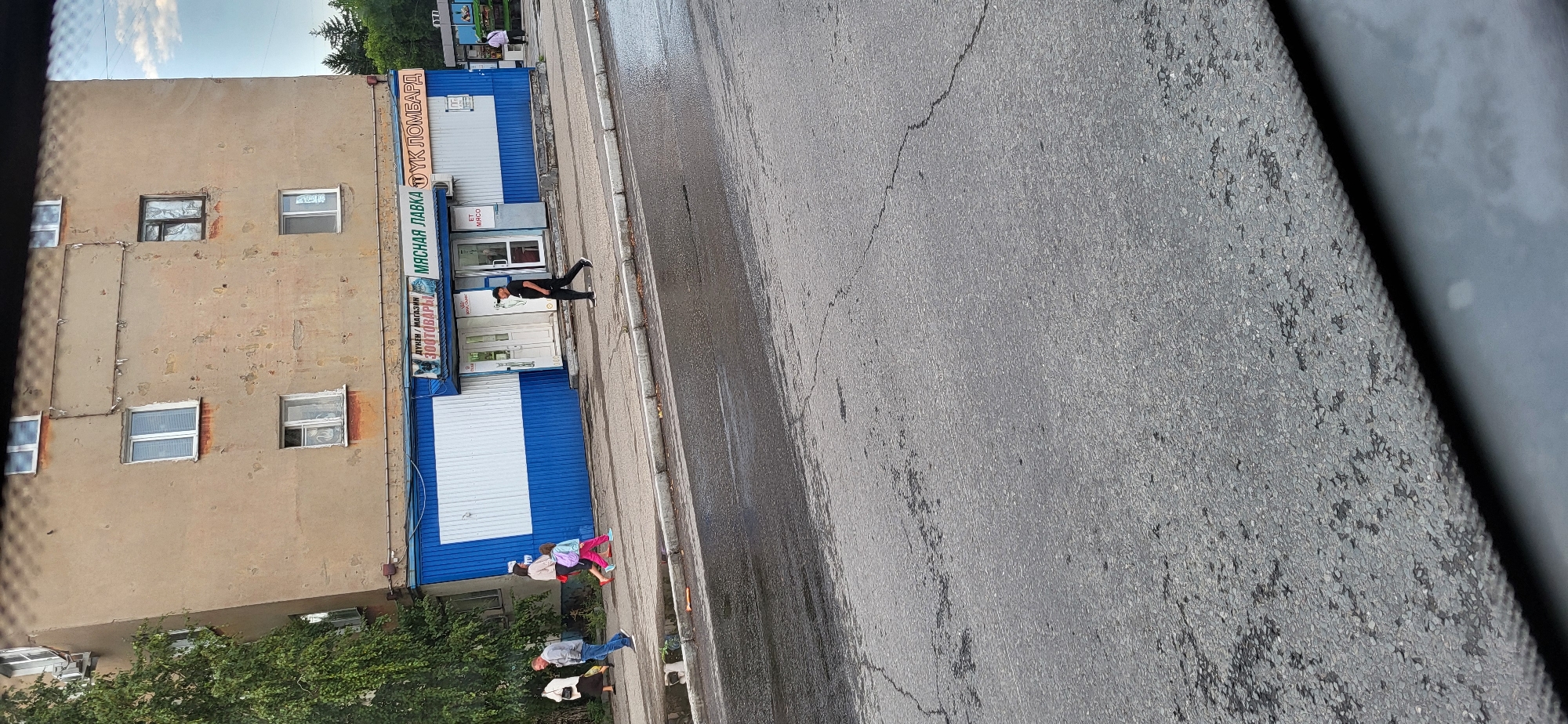 The image shows a street scene in Oskemen, Kazakhstan, in front of a dilapidated apartment building with a blue and white storefront. Several people are walking on the sidewalk. To the left, a woman in a dark dress and a man in a light jacket and jeans are walking away from the camera. Next to them, a woman in a pink top and pants, holding a pink backpack, walks with a young girl in a pink outfit. A man in a black shirt and pants walks towards the camera, crossing the street. The storefront appears to house a pet store and a meat shop, indicated by signage. The street in the foreground is wet, suggesting recent rain. The building has visible signs of wear and tear, with peeling paint and stained walls. The sky is overcast with some clouds.