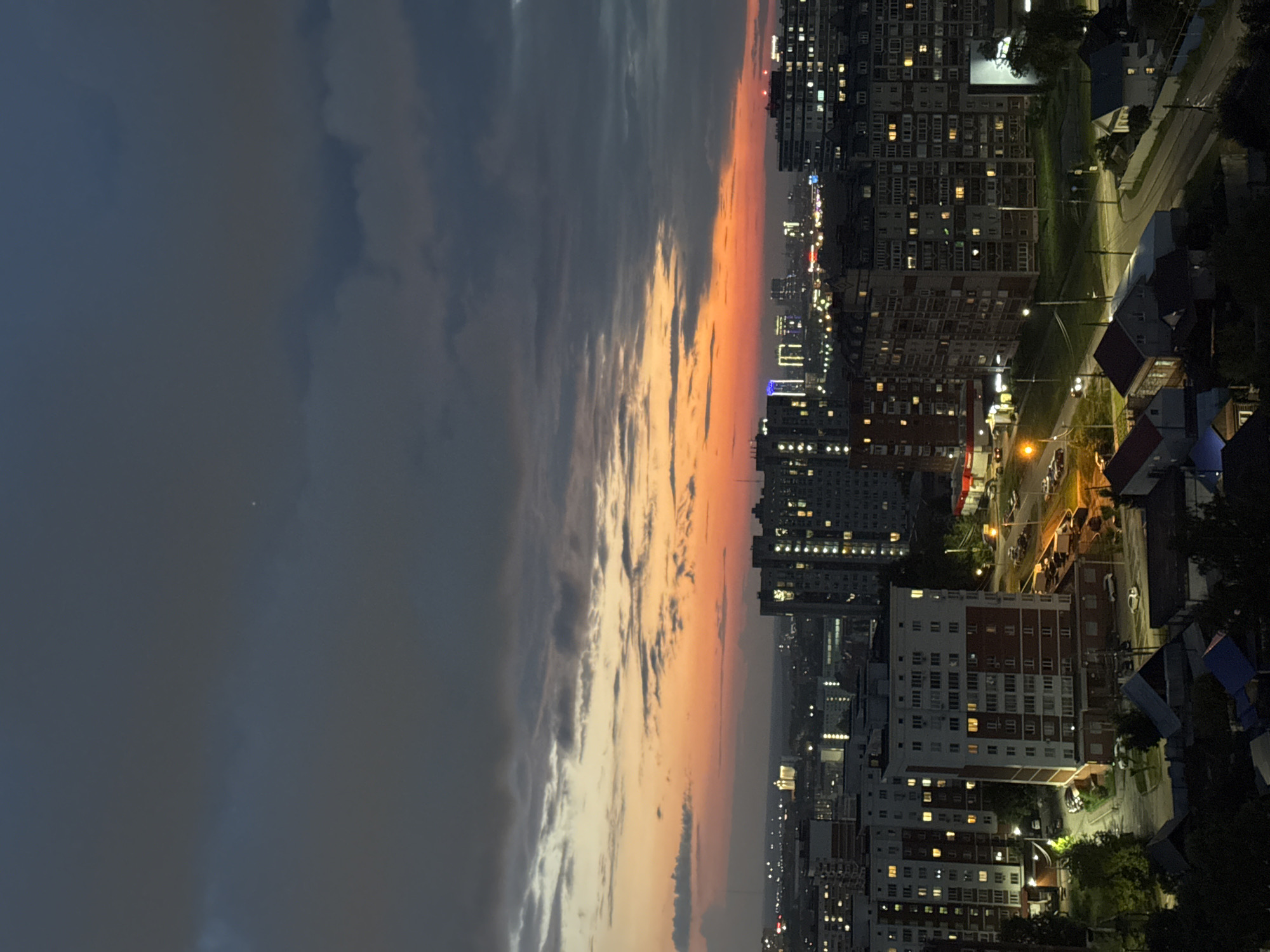 This is an aerial, vertical shot of an urban landscape at dusk in Izhevsk, Russia. The sky transitions from dark gray clouds at the top to a vibrant orange and pink sunset near the horizon. Several apartment buildings are visible, with lights on in many windows, indicating occupancy. Some buildings appear to be older, Soviet-era concrete structures, while others are more modern. Roads with streetlights and a few cars are present, along with some green spaces and trees. The overall scene depicts a residential area of the city as night begins to fall.