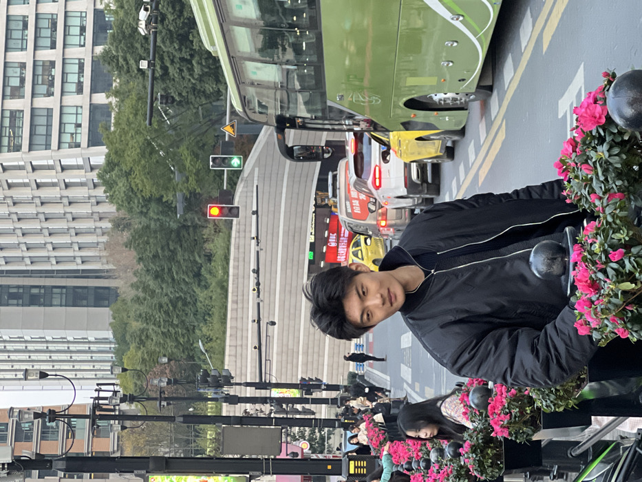 An East Asian male, approximately 20-30 years old, with short dark hair, is positioned in the immediate foreground, looking directly at the viewer with a slight smile. He is wearing a black bomber jacket over a dark collared shirt or sweater. He stands behind a series of dark rectangular planters containing vibrant pink flowering plants, possibly azaleas, which line the edge of a pedestrian area.
The scene depicts an urban street in Yuzhong District, China, during daylight hours under an overcast sky. To the right of the male subject, a multi-lane road is visible with queued vehicular traffic. A large green public transit bus occupies the rightmost lane. Behind it, a yellow taxi, a white SUV, and a dark sedan are stopped. Traffic lights in the midground show a red signal for straight traffic and a green arrow indicating a right turn. Overhead, multiple streetlights and traffic signal poles are present.
On the left side, beyond the planters, a sidewalk extends into the background. Several pedestrians are visible on the sidewalk, some appearing to walk past shopfronts. A female pedestrian is partially visible in the lower-left corner, positioned behind the planters.
In the background, modern high-rise buildings with glass and concrete facades dominate the skyline. A dense treeline is situated between the street level and the base of these buildings, indicative of either an elevated green space or an embankment. A concrete structure, possibly an underpass or tunnel entrance, is visible beneath the traffic lights.