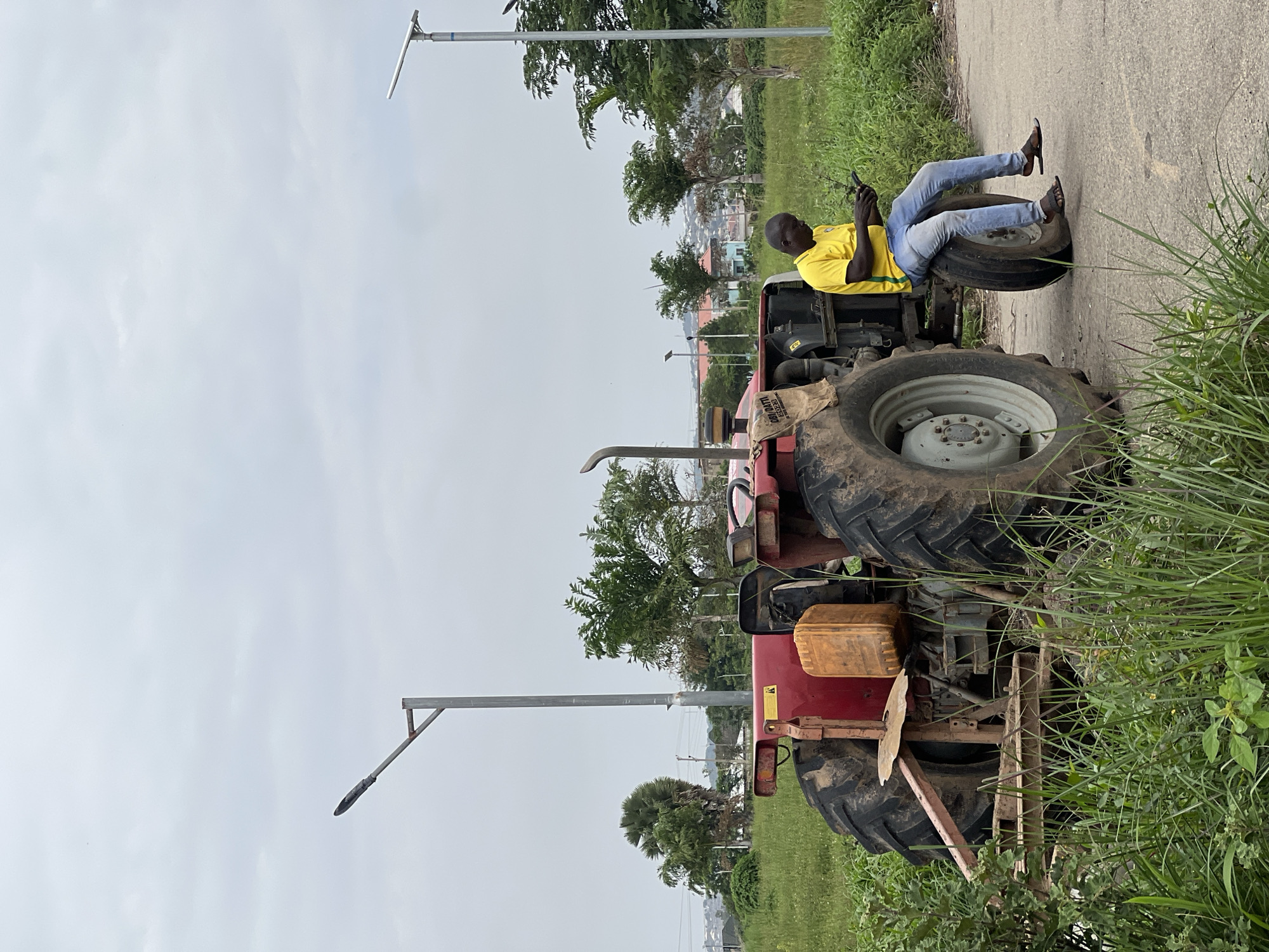 A Black man, wearing a yellow t-shirt with green stripes and blue jeans, is sitting on the rear tire of a red tractor. He is looking down at a black object in his hands, possibly a phone. The tractor is parked on an asphalt road adjacent to a grassy area. In the background, there are trees, utility poles with lights, and buildings visible in the distance. The sky is overcast. The scene appears to be in an outdoor, semi-rural or suburban area, potentially on the outskirts of Jos, Nigeria.