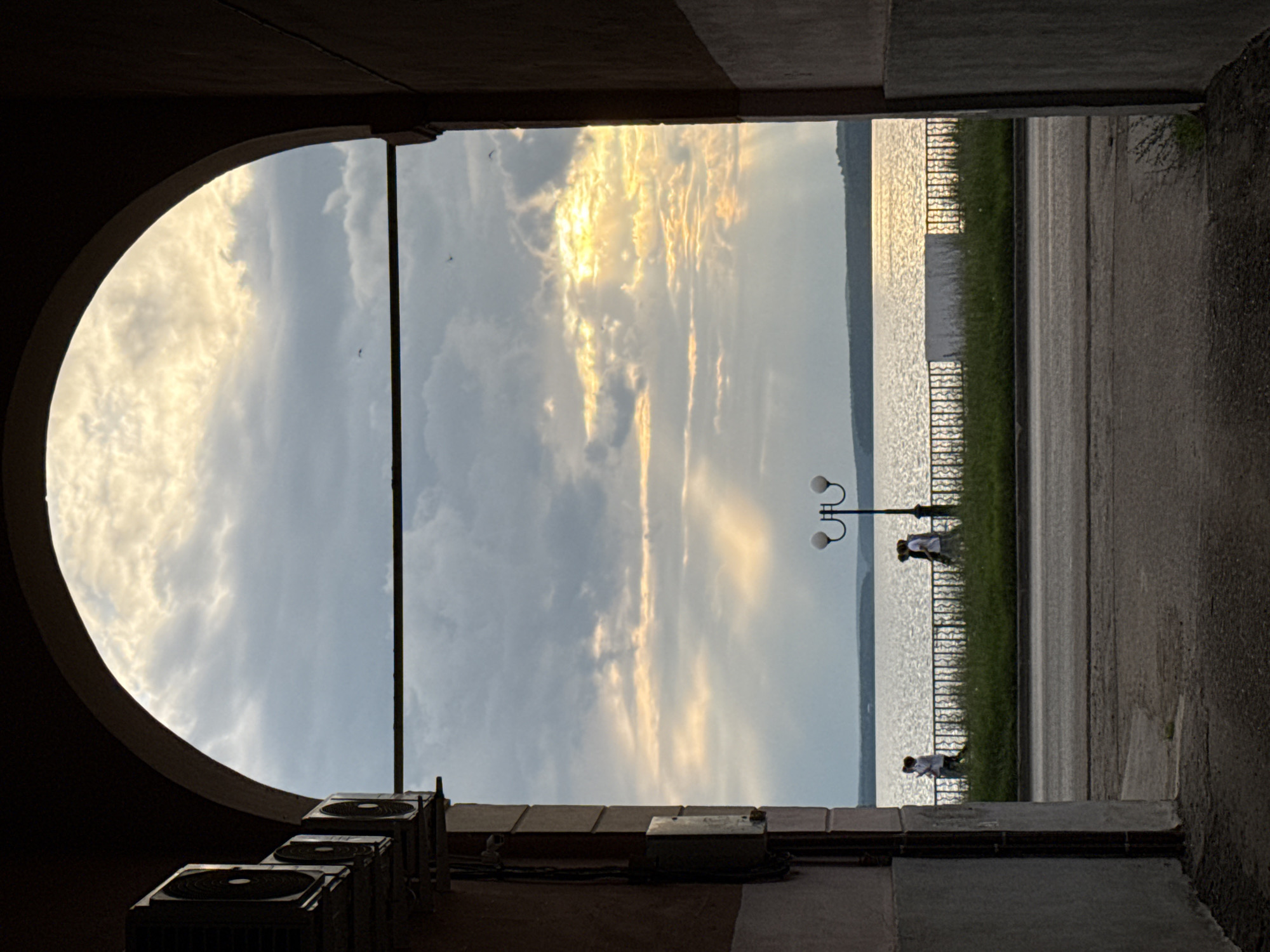 The image is a rotated, horizontal shot taken from within a dark, arched entryway. Through the arch, a bright sky filled with dramatic, golden-tinged clouds dominates the view. The sun appears to be either rising or setting, casting a warm glow across the sky and reflecting off a wide body of water in the distance. A faint shoreline with a grassy embankment and a decorative fence is visible. A street runs parallel to the water.
In the middle ground, a black lamppost with two white globes stands beside the fence. Two figures, likely people, are visible near the lamppost, appearing to stand or walk along the water's edge. They are wearing light-colored clothing. Further along the water's edge, another lone figure in light clothing is also visible.
In the foreground, at the bottom of the frame, are several large, dark air conditioning units mounted on a wall. The surrounding architecture suggests an urban or semi-urban setting. The overall mood is serene and picturesque, with the dramatic sky and water being the focal points. The time of day is indicated by the intense, warm lighting, suggesting either dawn or dusk. There is no visible text in the image. The location is Votkinsk, Russia.