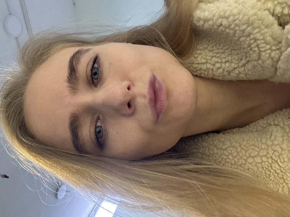 A light-skinned woman with blonde hair and blue eyes gazes into the camera. She wears a fuzzy, cream-colored jacket. Her eyebrows are thick and well-defined. Her lips are full and slightly parted. The photo appears to be taken from a low angle, giving a slightly distorted perspective of her face. A portion of the ceiling with a light fixture is visible in the background.