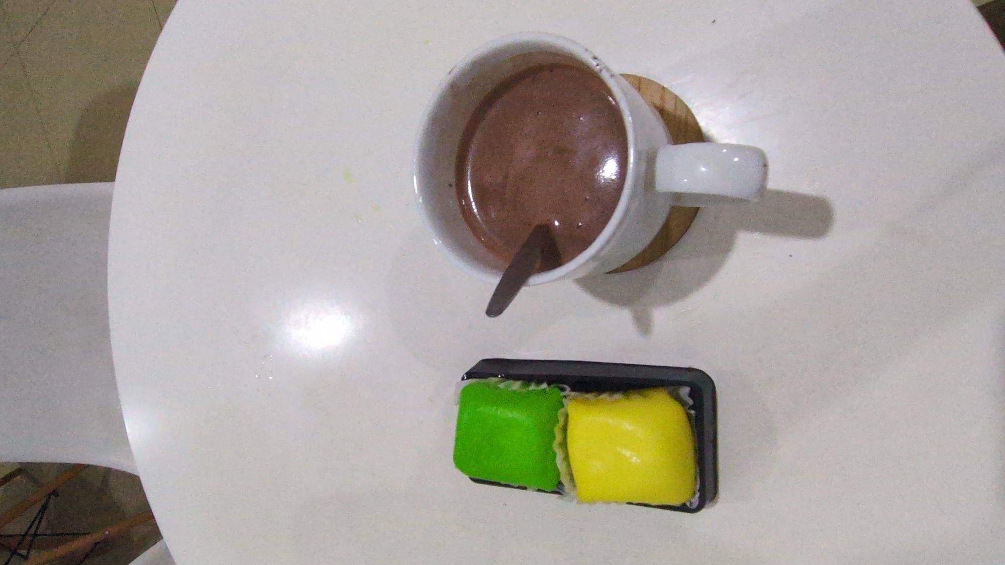 A top-down view shows a white table with a cup of chocolate drink, a spoon, and a small container holding green and yellow sweets. The cup sits on a wooden coaster. A white chair is visible in the lower left of the frame. The location is likely a cafe or similar establishment in Phan Rang – Tháp Chàm, Vietnam.