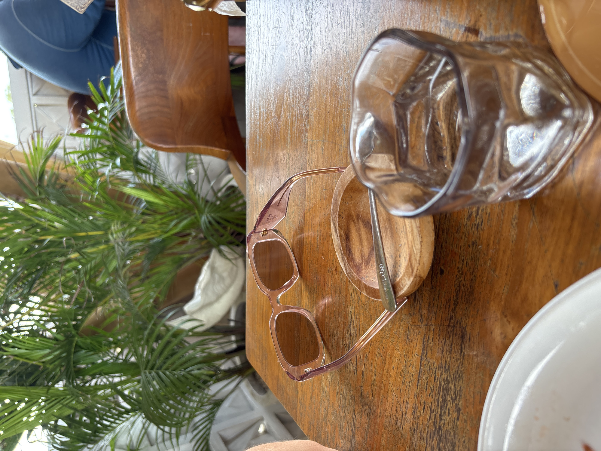 The image depicts a sunlit, casual setting, likely a cafe or restaurant in Kuta Selatan, Indonesia. The main focus is a rustic wooden table, viewed from a close-up, slightly overhead perspective.
On the table, a pair of pink-tinted, square-framed sunglasses are prominently displayed. The brand "PRADA" is visibly engraved on the inner temple of the sunglasses. Adjacent to the sunglasses, a small, light-colored wooden bowl holds a slender metal stirring stick, which has some indistinct text, possibly "COZY," etched onto it. To the right, an empty, clear glass with a faceted design sits on the table. A portion of a white plate is visible in the bottom right corner. The wooden table exhibits a natural grain and some light scratches, suggesting a well-used but inviting surface.
In the background, out of focus, are vibrant green palm-like plants, indicating a tropical or garden setting. Beyond the foliage, a wooden chair back and a person dressed in or seated on blue fabric are partially visible. Bright natural light illuminates the scene, suggesting it is daytime. The overall impression is one of a relaxed and pleasant environment.