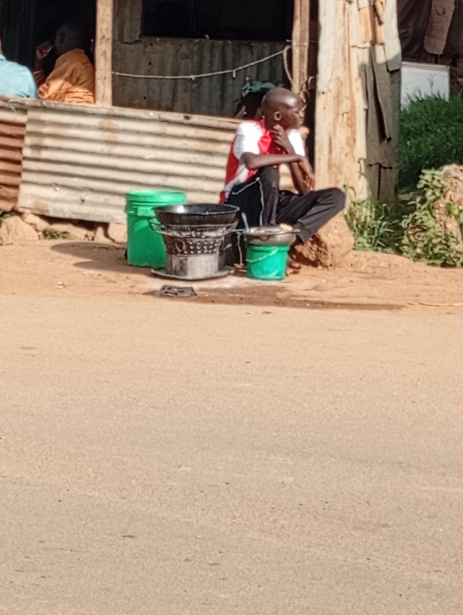 In Rafin Par, Nigeria, a young Black male is seated on the ground, facing right, with his knees bent and his right leg tucked under him. He wears a white t-shirt with red trim around the neck and arms, and black pants with white stripes. His left arm rests on his knee, and his right hand is near his chin, as if in thought. He is positioned in front of a makeshift stall constructed from corrugated metal and wooden poles. Next to him are two green buckets, one larger than the other, and a metal cooking brazier with a large black pot on top. Behind him, a figure in an orange garment is visible inside the stall. The ground is dirt and sand, with some scattered rocks and dry vegetation. In the background, there is more corrugated metal fencing and some green foliage. The scene appears to be an outdoor marketplace or roadside setup.