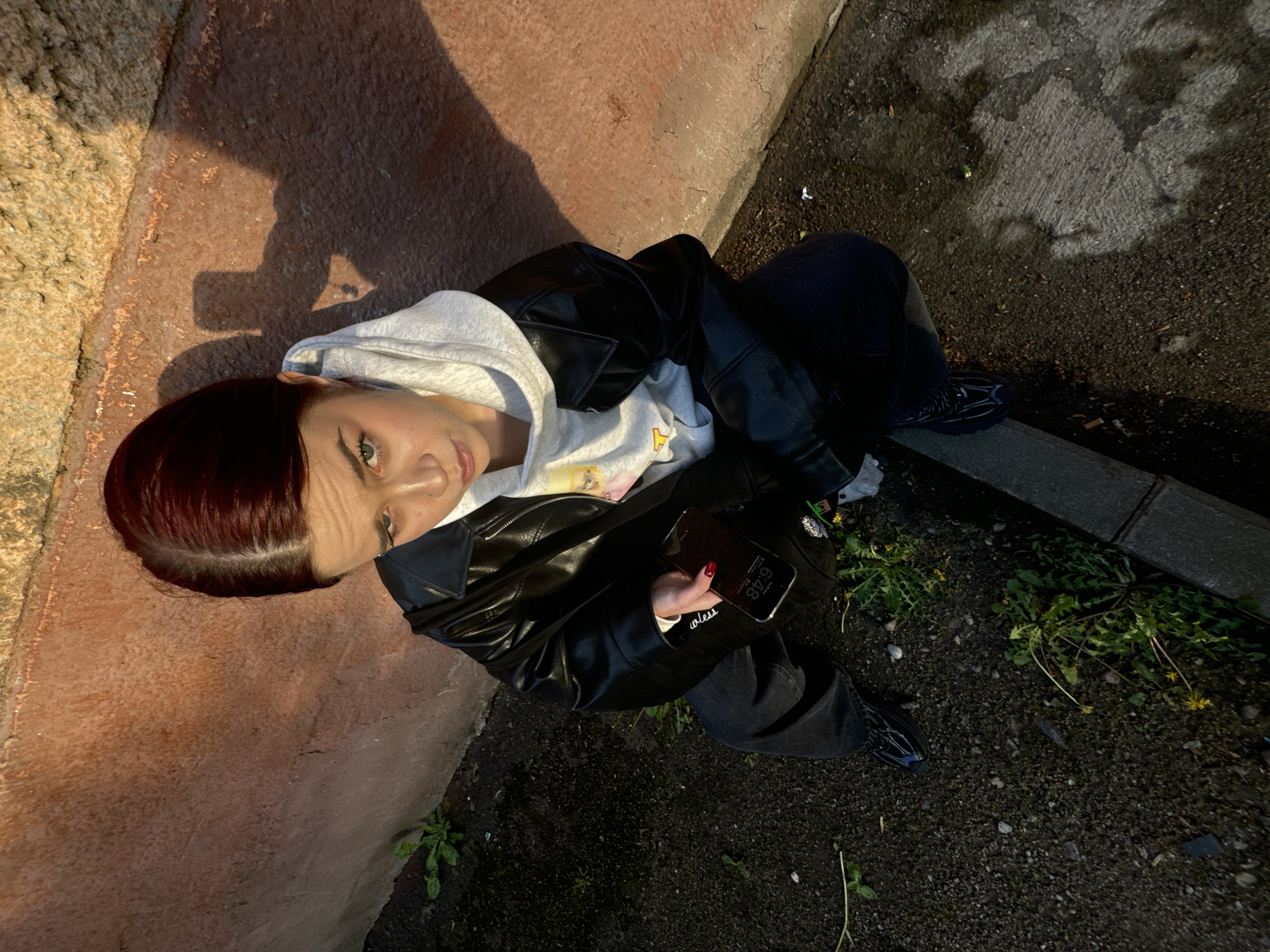A high-angle shot shows a woman sitting on the edge of a concrete structure, possibly a raised planter or wall, next to an asphalt surface. The woman has auburn hair and fair skin, wearing a light grey hoodie with a graphic and a black leather jacket. She is holding a smartphone in her right hand, displaying a time of 6:48. She appears to be looking up at the camera. The concrete structure is pinkish-beige, with a shadow cast across it. The asphalt is dark and slightly damp, with scattered small plants growing in the cracks and at the edge of the concrete. The location is urban, with a curb visible at the edge of the asphalt.