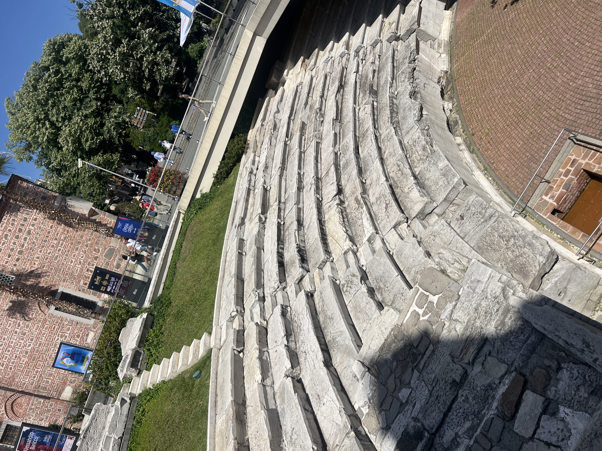 The image depicts the ancient Roman theatre in Plovdiv, Bulgaria. The theatre's stone seating tiers are arranged in a semi-circle, showing signs of wear and age. Grass grows between the seating sections. A modern walkway with a railing is visible above the theater. A brick building and a tree are located to the left of the theater. Posters and people are visible near the brick building. A brick-paved area is on the right side of the theater.