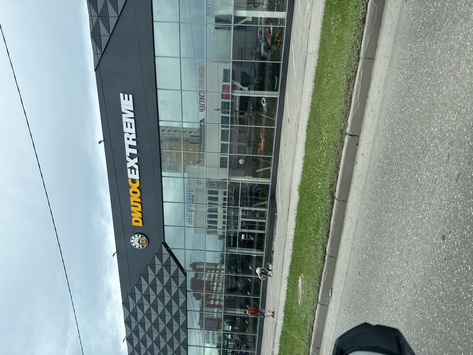 The image captures the exterior of a business with large glass windows and a prominent sign reading "ДИЛОС EXTREME" in yellow lettering. The building has a modern architectural design with a dark gray facade and a geometric pattern on one section. Reflections of nearby buildings and the sky are visible on the glass.
In the foreground, there is a paved area with grass and a curb. Two individuals are present outside the building. One person, wearing a red backpack and shorts, stands looking towards the building. Another person, in the process of bending down, appears to be interacting with something on the ground near the entrance. The area around them suggests an outdoor commercial setting. The scene is viewed from a slightly elevated perspective, likely from a vehicle. The overall environment suggests an urban or suburban commercial district.