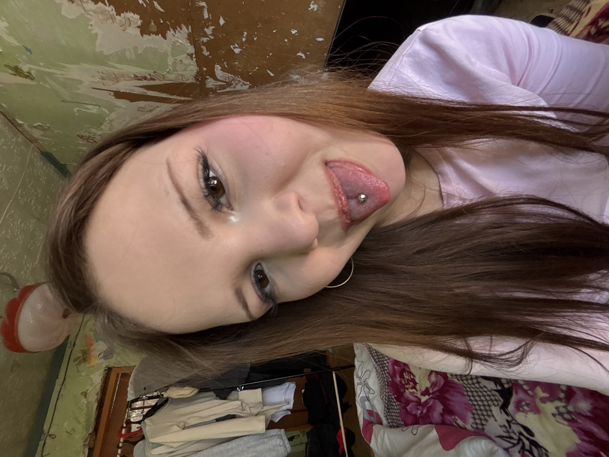 A woman with long brown hair and a tongue piercing is posing for a close-up selfie. She is wearing a light pink shirt and a small silver hoop earring. The background appears to be a bedroom interior. The walls have peeling paint, and a clothing rack with various garments is visible. Part of a floral-patterned comforter can also be seen. The image was not taken in Фоки, Russia.