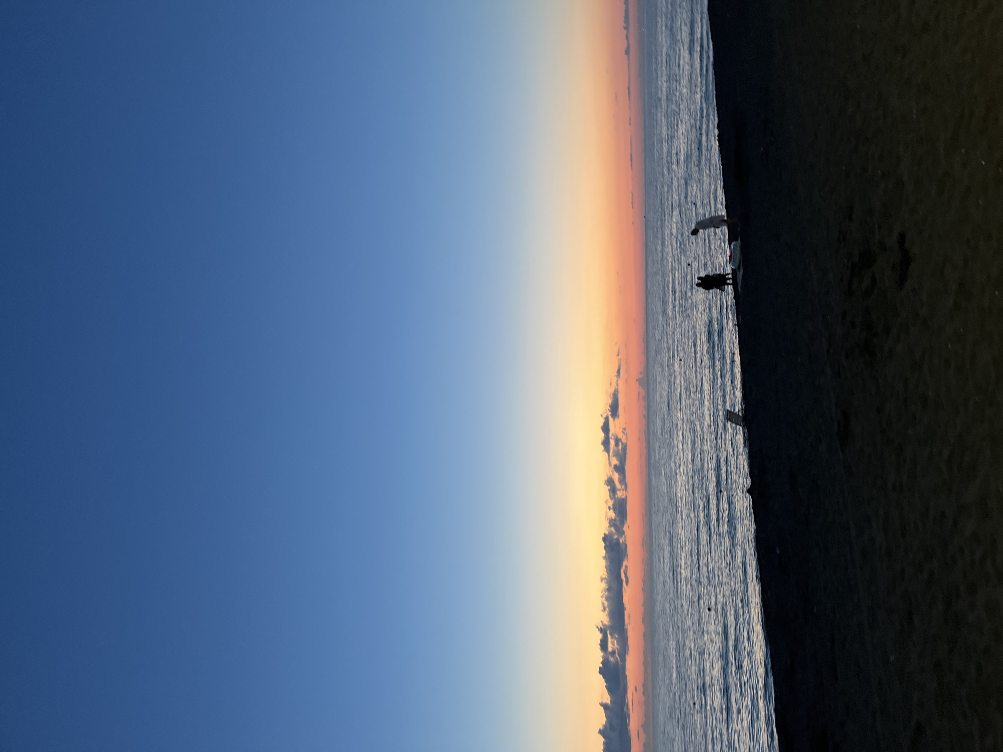 The image depicts a serene beach scene at sunset in Ureki, Georgia. The sky transitions from a deep blue at the top to a vibrant gradient of orange and pink hues near the horizon, with a line of dark clouds silhouetted against the colorful sky. The sea is calm, with gentle waves lapping against the dark sand beach. In the foreground, three figures are visible on the beach. Two figures stand close together, appearing to be engaged in conversation, while a third figure stands slightly apart, facing the water. The sand itself appears dark and perhaps wet from the receding tide. In the mid-ground, a white object, possibly an inflatable or a beach chair, is situated near the water's edge. The overall impression is one of tranquility and the quiet beauty of a coastal evening.
