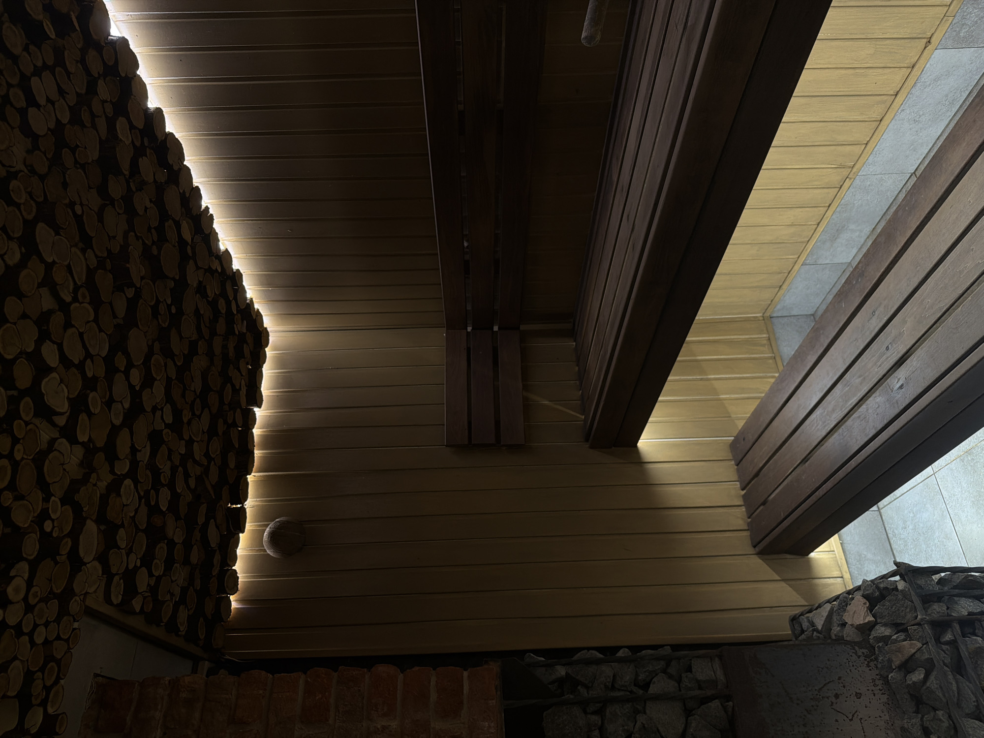 The image depicts the interior of a building, potentially a sauna or similar space, in what is stated to be Kamennoye, Russia. The walls appear to be constructed of horizontal wooden planks, with some areas darker and others lighter, suggesting different types of wood or finishes. One section of the wall is decorated with a dense arrangement of small, round wood pieces, possibly log slices, creating a textured effect. Adjacent to this, there is a brick structure, possibly a stove or fireplace base. In the lower right, there is a section of wall with a stone covering held in place by metal wiring. The lighting is dim, with a light source illuminating the edge of the log wall, accentuating its texture. Above, there are structural wooden beams. The overall atmosphere is rustic and dimly lit.