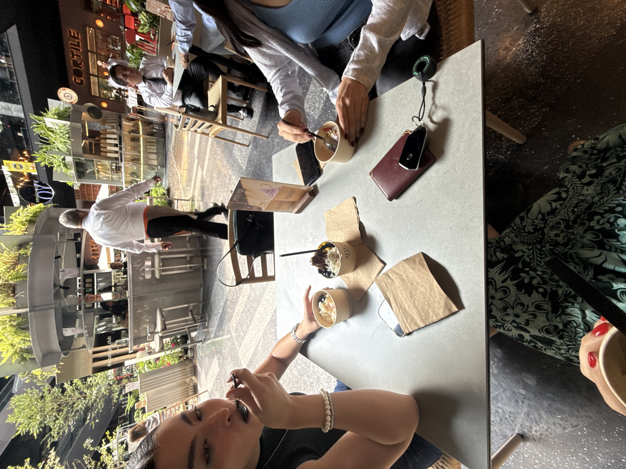 The image shows an overhead view of a table at a restaurant in Mexico City, Mexico. Three people are partially visible. One woman is facing the camera, holding a utensil, with a beaded bracelet on her wrist. Another person is across the table, holding a spoon and eating from a cup. A third person in a floral-patterned dress is seated to the right. Several cups of food are on the table with paper bags. Keys and a wallet are next to the cup. Other patrons and restaurant staff can be seen in the background. The restaurant is called "Cortile the coffee club".