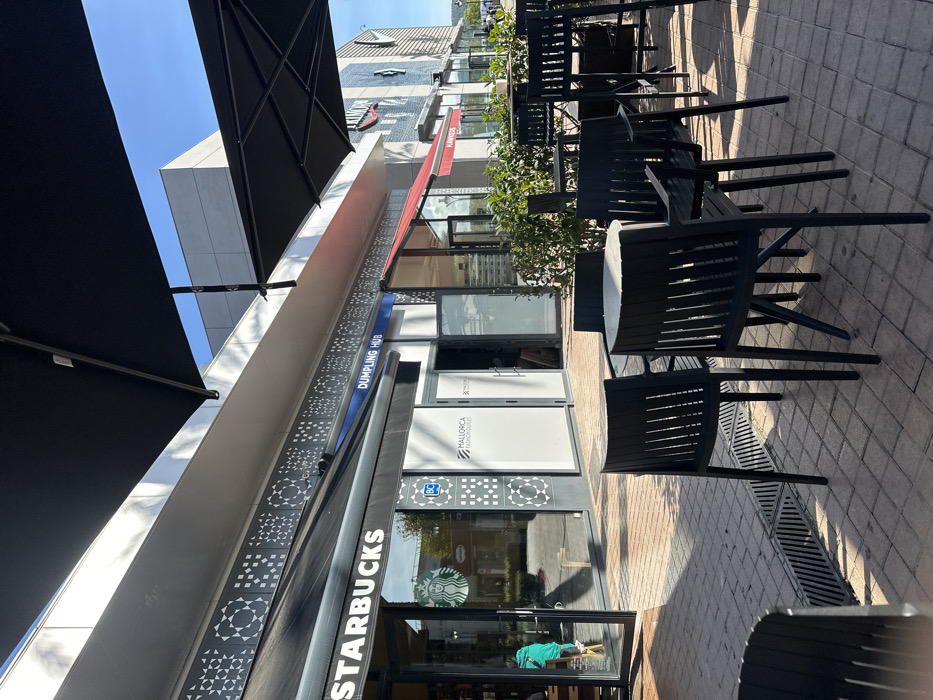 The image displays an outdoor paved plaza with seating in front of several retail storefronts at Mallorca Fashion Outlet in Marratxí, Spain, under a clear blue sky.
In the foreground, a light-colored rectangular paver stone plaza features a linear drainage grate on the left. Centrally, a round table with a light-colored surface is surrounded by four black outdoor chairs, each with slatted backs and armrests. To the right, additional black outdoor tables and chairs, including high-top configurations, are partially obscured by low, green ornamental bushes planted in rectangular beds. Strong, diagonal shadows from the left extend across the paving and seating area, indicating direct sunlight.
The midground consists of a row of storefronts. On the far left, a Starbucks coffee shop is visible, featuring large glass windows, a dark awning with "STARBUCKS" in white lettering, and the green siren logo on the glass. A person wearing a green top is partially visible inside the Starbucks near its open glass entrance door. Adjacent to Starbucks, two storefronts are labeled "MALLORCA FASHION OUTLET" with white signage on dark grey backgrounds; their glass facades are covered with white opaque material. One of these units has an open door, revealing a dimly lit interior with some visible wooden structures. Above these two units, a blue sign reads "DUMPLING HUB." Further to the right, a red awning and a sign for "HAWKERS" are visible. The building facade above the storefronts is light-colored, featuring a grey decorative panel with repeating geometric square and circular patterns. Large black cantilevered umbrellas extend over the seating area, providing shade.
The background reveals sections of a modern building with clean, light-colored paneling against the clear blue sky, and distant green foliage to the far right.