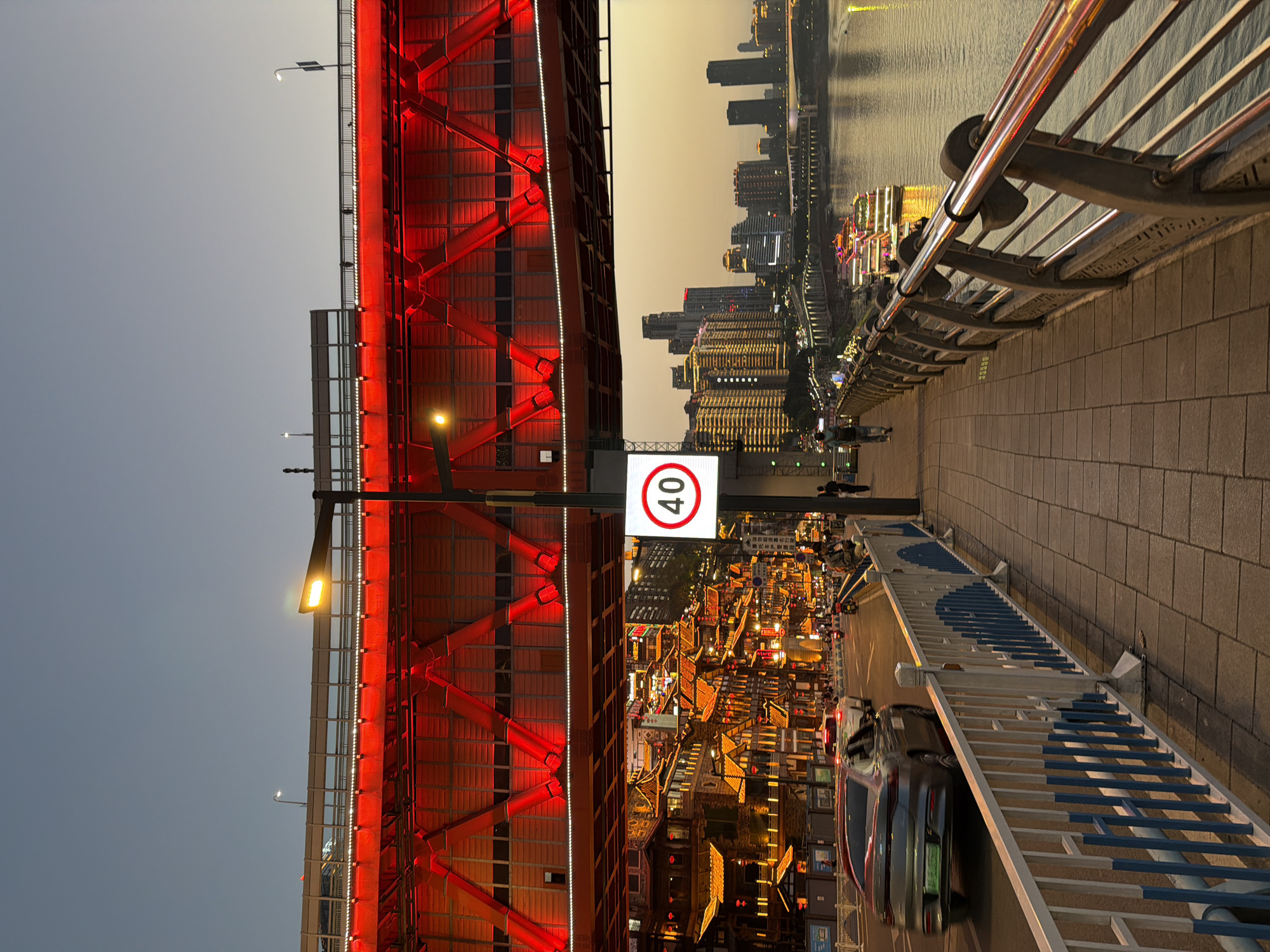 The image depicts a scene in Yuzhong District, Chongqing, China, featuring the Hongyadong commercial complex illuminated at dusk. A bright red bridge spans the foreground, marked with a "40" speed limit sign. Below the bridge, the multi-tiered traditional architecture of Hongyadong glows with warm light. Beyond, skyscrapers line the riverfront, their lights reflecting on the water. Pedestrians are walking on the sidewalk next to a blue and white safety barrier with a silver railing. Cars are on the road next to the sidewalk.