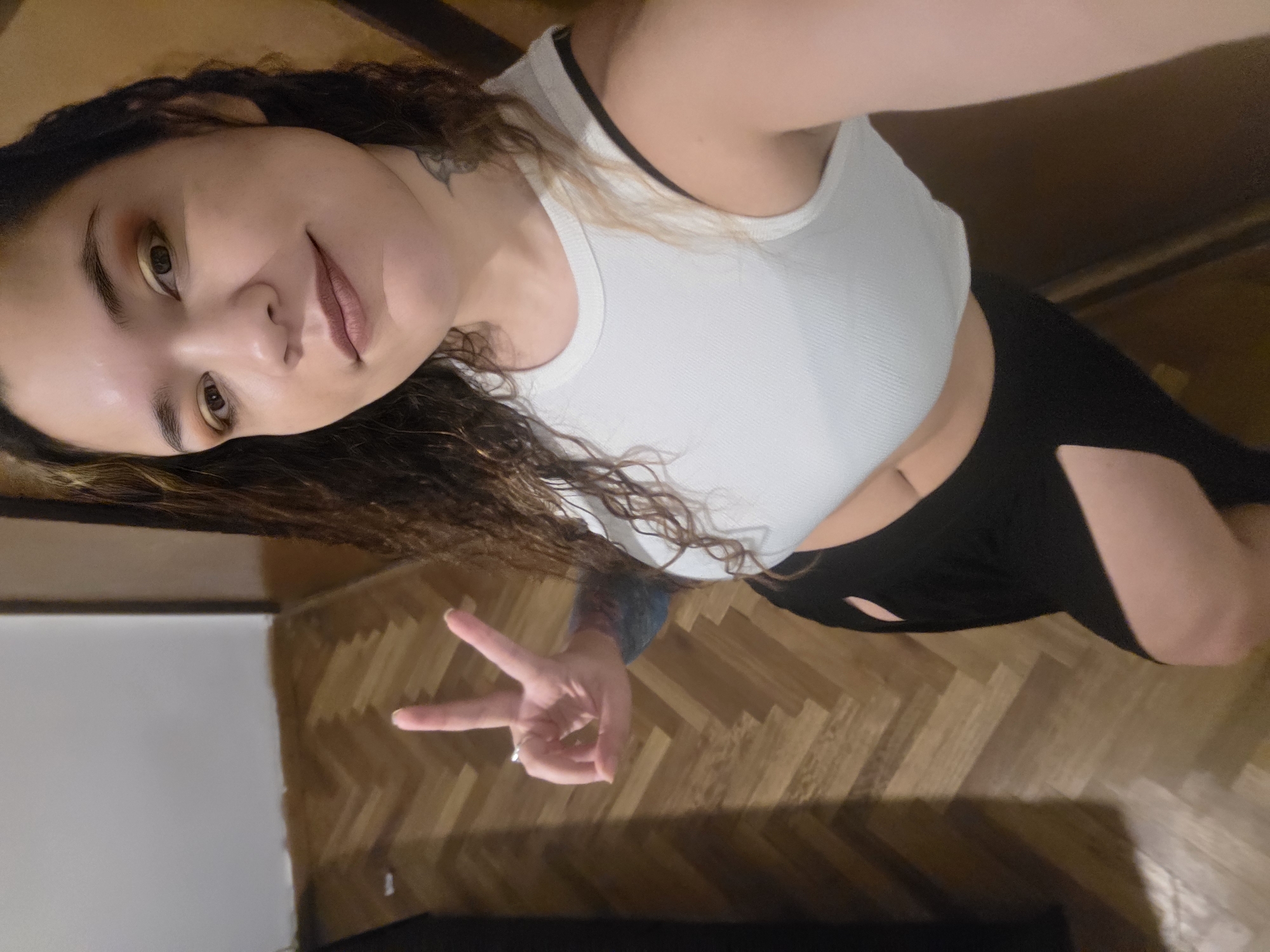 The image is a self-portrait (selfie) of a person, rotated 90 degrees clockwise. The subject is a young-looking individual with long, wavy to curly dark brown hair, with some lighter, possibly blondish, streaks visible, especially on the side of their head. They are looking directly at the camera with a gentle, slight smile on their face. Their eyes are dark, and they are wearing eye makeup that includes a shimmering gold or bronze eyeshadow. Their lips have a natural-toned lipstick.
The person is wearing a white, ribbed, sleeveless crop top, revealing a portion of their midriff. A black strap, likely from a bra or another undergarment, is visible beneath the white top near the shoulder. On their lower half, they are wearing black bottoms, which appear to be shorts or leggings, with a distinctive cut-out design on the side of one thigh/leg.
Their right hand (left side of the image as oriented, but right hand of the person) is extended towards the camera, making a "peace" sign (V-sign) with their index and middle fingers. A ring is visible on the ring finger of this hand. A dark tattoo is partially visible on their right shoulder/upper arm, peeking from under their hair. Another tattoo, with blueish tones, is visible on their left forearm.
The background suggests an indoor setting. The flooring is made of wooden planks arranged in a herringbone pattern. A plain, light-colored wall is visible in the background. The lighting appears to be warm and soft, possibly from indoor artificial light sources. There is no visible text in the image.
