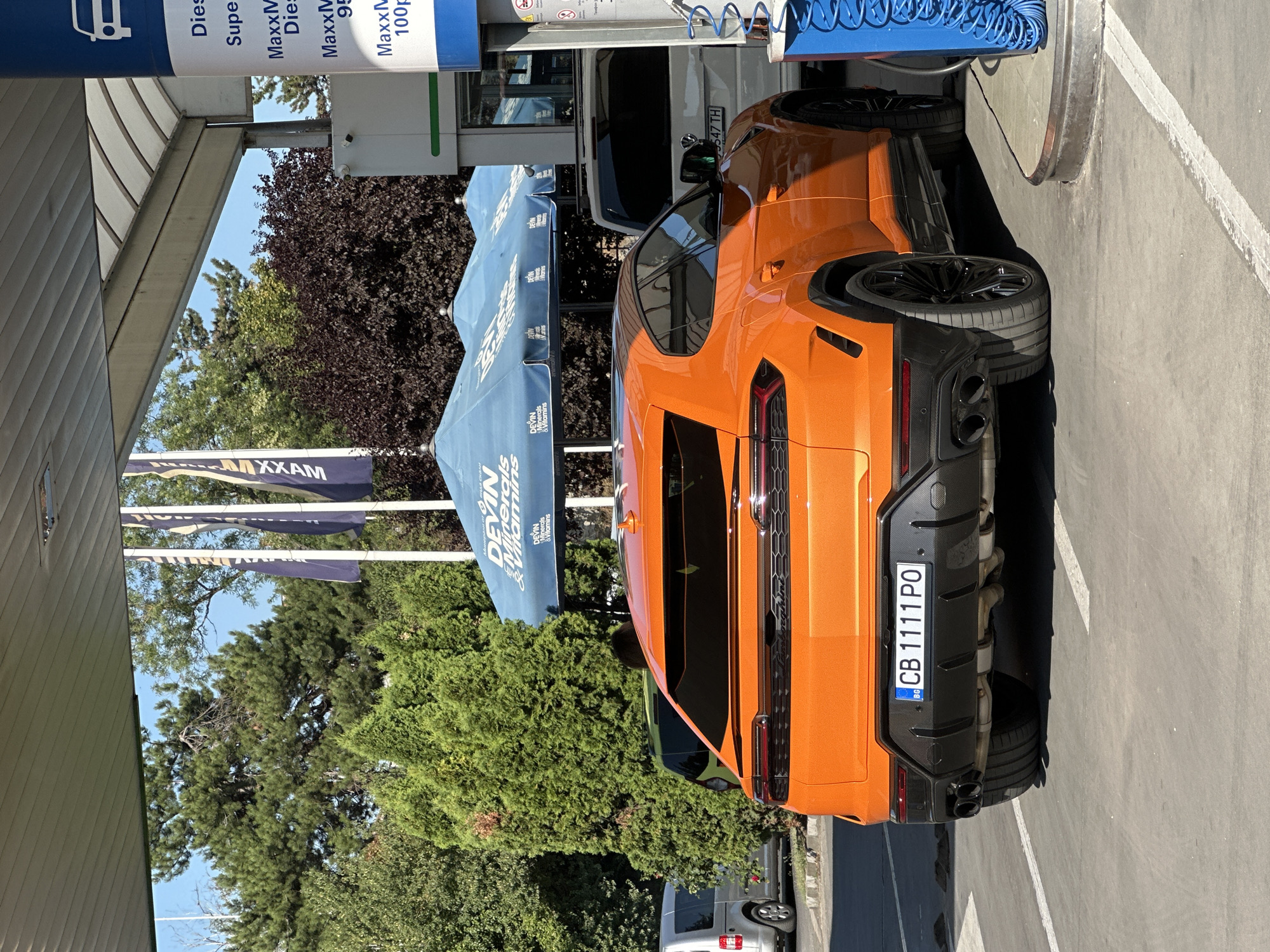 This is a vertical, eye-level, medium shot of an orange Lamborghini Urus parked at a gas station on a sunny day in Veliko Tarnovo, Bulgaria. The car is the primary subject, taking up a large portion of the frame, and is viewed from the rear three-quarters angle. Its bright orange paint sparkles in the sunlight, highlighting its aggressive design, with prominent black accents on the diffuser and rear bumper. The car's license plate, "CB 1111 PO," is clearly visible.
In the background, to the left, is a gas pump canopy with signage indicating fuel types like "Diesel" and "MaxxM" with prices. Blue awnings with the "DEVIN" logo, promoting minerals and vitamins, are also visible. Further back, lush green trees and a dark, leafy tree provide a natural backdrop, contrasting with the man-made structures of the gas station. A white van with a partial license plate, "9547 TH," is parked behind the Lamborghini, suggesting a busy or functional area. The overall scene conveys a sense of everyday activity at a gas station, with a luxury vehicle as the focal point. The bright sunlight and clear blue sky suggest pleasant weather conditions. The image is rich with brand names and specific car details, indicating a focus on commercial products and automotive aesthetics.
