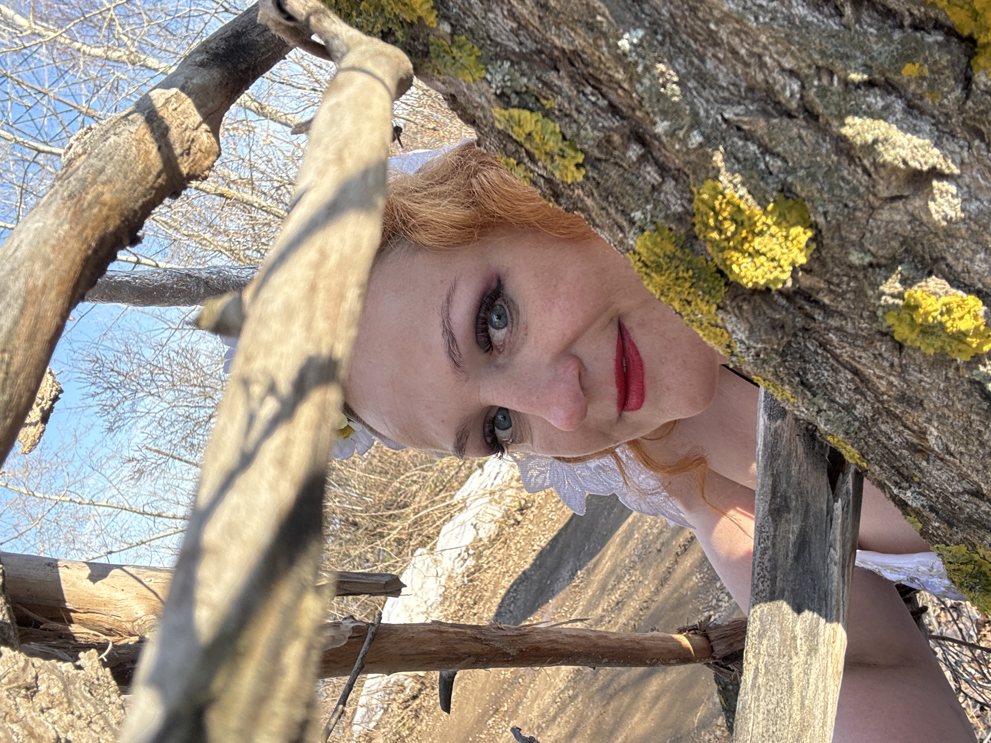 The image depicts a woman with red hair, blue eyes, and red lipstick, framed by tree branches. The branches on the right have patches of yellow lichen. She is wearing a white lace garment. The background suggests an outdoor setting with a mix of vegetation and a dirt path. The image is taken at an angle, with a visible patch of blue sky and bare tree branches in the upper left. Based on the prompt, this location is in Chaikovsky, Russia, although this cannot be verified from the visual information alone.
