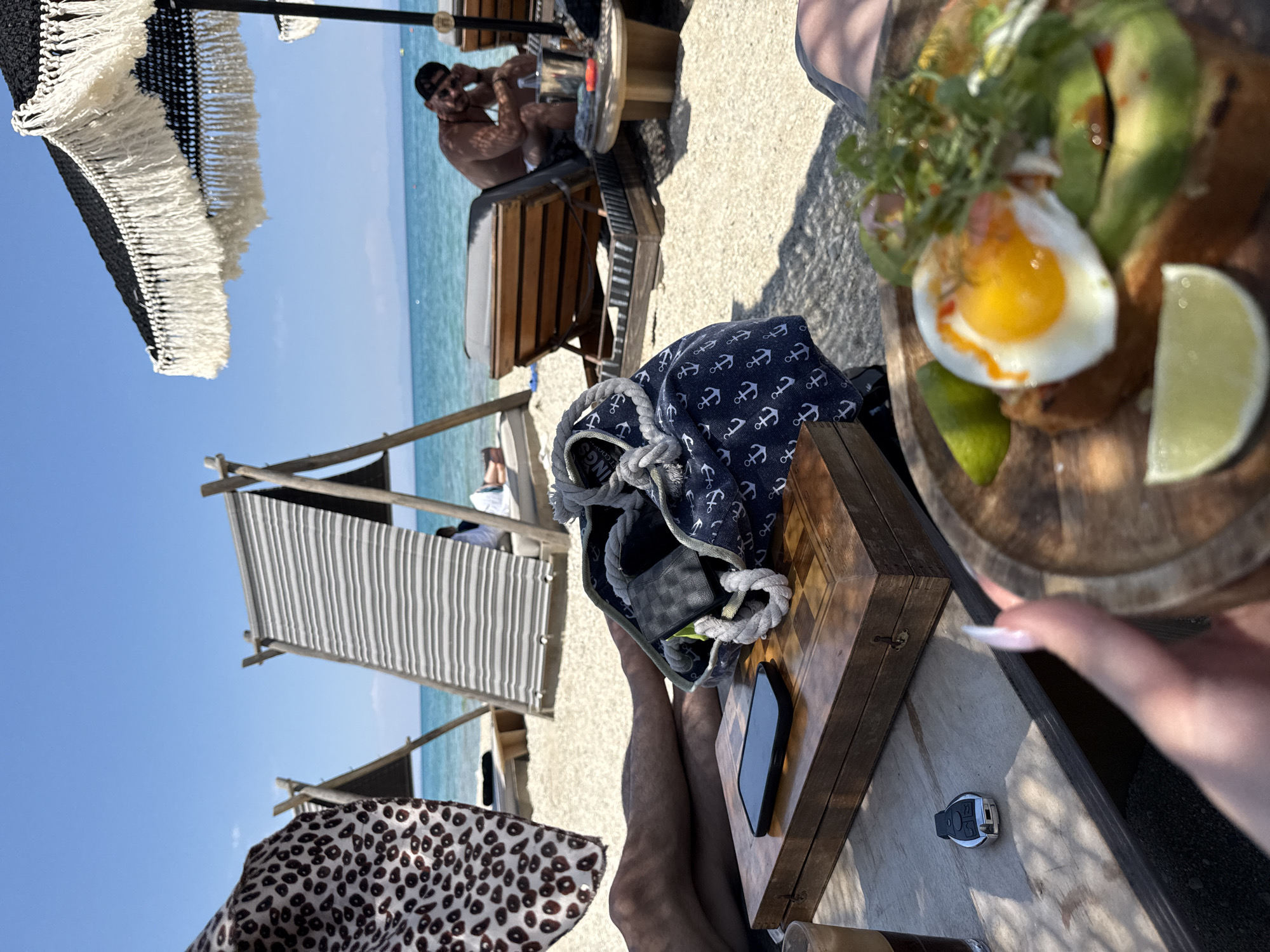 The image depicts a sunny day at a beach in Pallini Municipal Unit, Greece. In the foreground, a person's legs are visible, along with a wooden table. On the table, there's a mobile phone, a car key fob, and a small wooden board with a meal. The meal includes a poached egg, avocado slices, greens, and lime wedges, presented on a wooden platter. A person's hand with manicured nails reaches towards the platter.
In the mid-ground, a navy blue bag with a white anchor pattern lies on the table, held by a thick rope. A beach umbrella with white fringe provides shade, and striped canopies are set up further back on the sandy beach. The turquoise sea stretches out under a clear blue sky.
In the background, a man is seated on a wooden lounger, looking towards the camera. He is wearing a black baseball cap and appears relaxed. The overall atmosphere suggests a leisurely day at the beach, likely during daylight hours given the bright sunlight and shadows. There are no discernible signs or text in the image.
