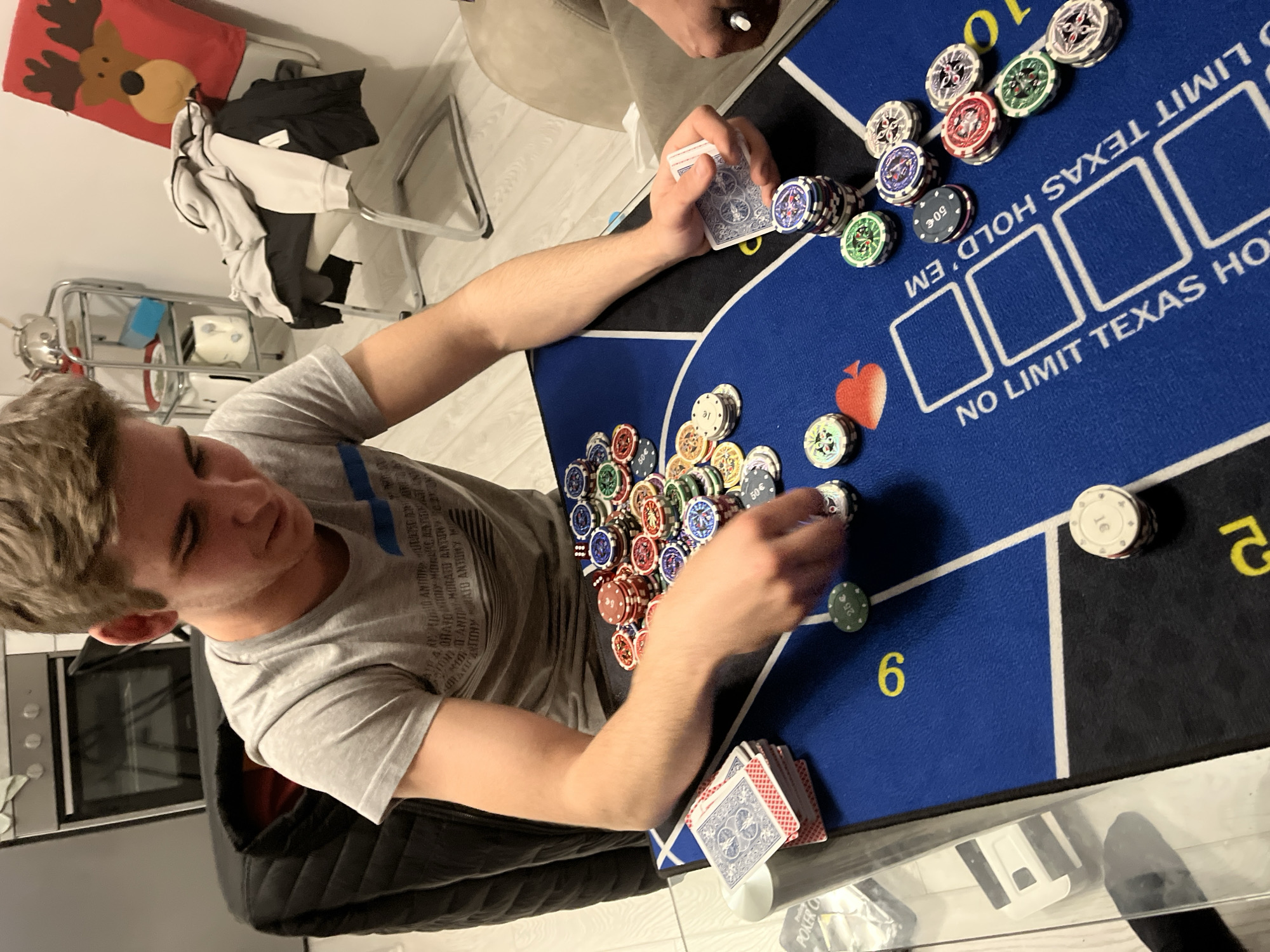 A young man with blond hair is playing Texas Hold'em poker on a blue felt mat with the words "NO LIMIT TEXAS HOLD'EM" printed on it. He is sitting at a table with poker chips and cards. He is wearing a gray t-shirt. There is a stack of chips in the center of the table, and another player is visible at the top of the image. A stack of cards sits at the bottom of the table. The setting seems to be indoors. The OCR data suggests "POKER C" is on the table and that "LIMIT TEXAS HOLD'EM" is printed on the mat. The OCR data includes "ANTONY" suggesting this person's name. There is no information to place the location of the scene in Burgas, Bulgaria.