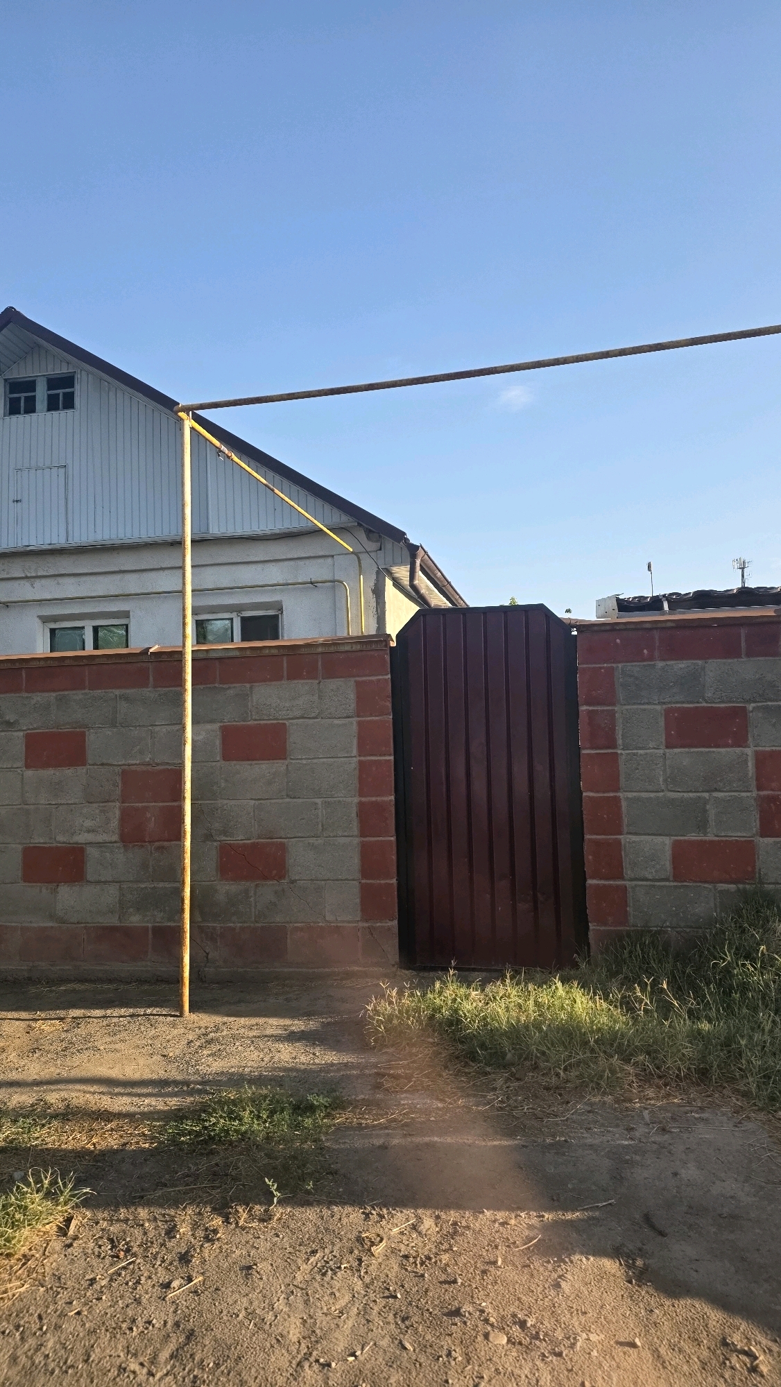 The image depicts a section of a brick and cinder block wall topped with a brown metal gate. The wall is constructed with alternating grey cinder blocks and red brick accents. A yellow metal pole, likely part of a clothesline or some other support structure, is positioned vertically in front of the wall and extends upwards towards a horizontal bar. Behind the wall, a portion of a white building with a dark roof and two windows is visible. The sky above is clear and blue. The foreground shows a dirt path with some patches of dry grass and shadows cast by the structures. The scene is likely located in Chuy, Kyrgyzstan, given the context.