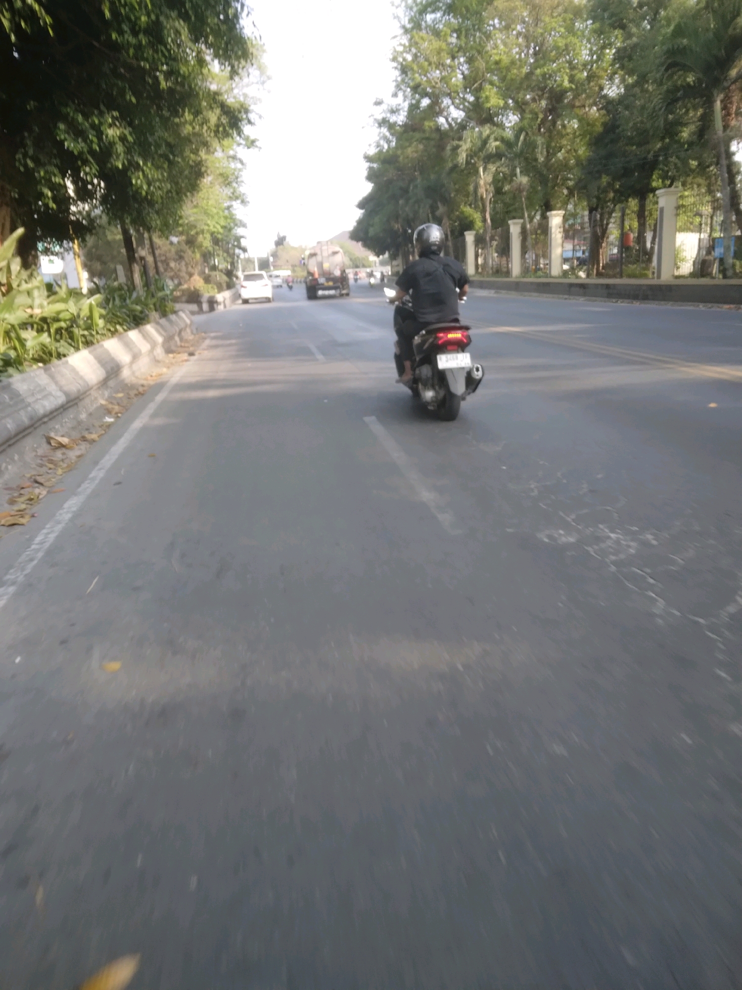 A low-angle, outdoor, point-of-view shot captures a person riding a dark-colored scooter down a paved road lined with trees and a concrete barrier on the left. The scooter rider, wearing a black shirt and helmet, is positioned in the center of the frame, moving away from the viewer. The road appears to be a multi-lane street with some faded white markings. In the background, a white car and a truck are visible, suggesting traffic. The sky is bright and overcast. The scene suggests a typical street in Sokaraja, Indonesia, during daylight.