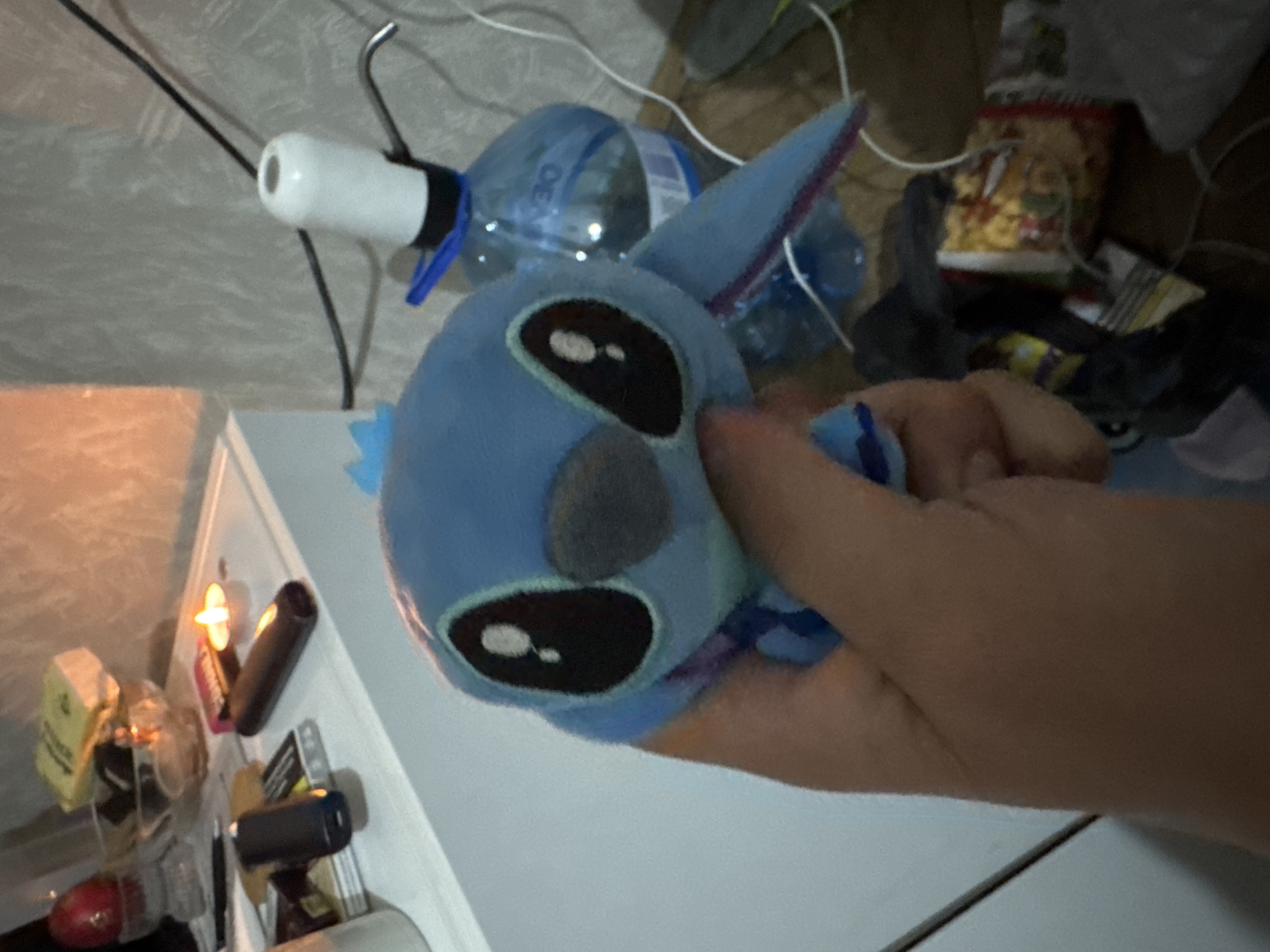 The image displays a close-up, rotated view of an adult's hand gently holding a blue plush toy, resembling Stitch from Lilo & Stitch. The toy features large black eyes with white highlights and a grey oval nose.
The scene is an indoor, somewhat cluttered environment with dim lighting, suggesting evening or night. On a light-colored surface to the left, various items are scattered, including containers, a yellow box, and two black cylindrical objects. One of these black objects emits a distinct orange glow. In the background, behind the plush toy, several plastic water bottles are visible; one clearly shows the brand "DEYA" on its label. Also present is a white and black device with a metallic hook or stand, connected to black wires, possibly a small lamp or charger. Further back, a red and gold snack package and loose white wires are discernible against a textured wall or surface.
The "DEYA" brand on the water bottle serves as a specific location cue, aligning with the context of Sofia, Bulgaria. The overall ambiance is casual and personal, typical of a domestic setting.