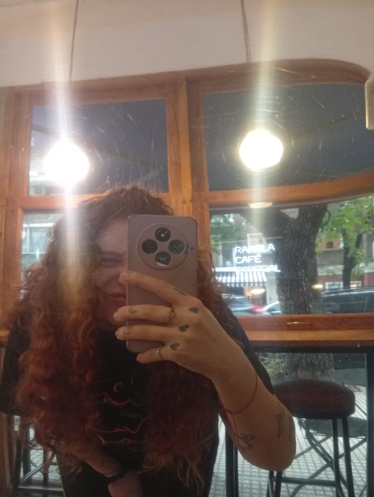 A woman with red curly hair is taking a selfie in a cafe. The phone is a lavender color with four lenses and a circular design. Her left hand, holding the phone, has tattoos of small hearts and a crescent moon. She wears a ring on one finger. She is wearing a black shirt. A red string bracelet is around her right wrist, and there is a tattoo on her right forearm.
Behind her, through a window, is a street scene. The words "RAMBLA CAFE" are visible on a window across the street. A dark-colored car is parked in front of the cafe. A large tree is in the background. The cafe appears to be in Buenos Aires, Argentina. A wooden-framed window and a hanging light fixture are visible in the cafe where the woman is. A bar stool is to the right of the woman.