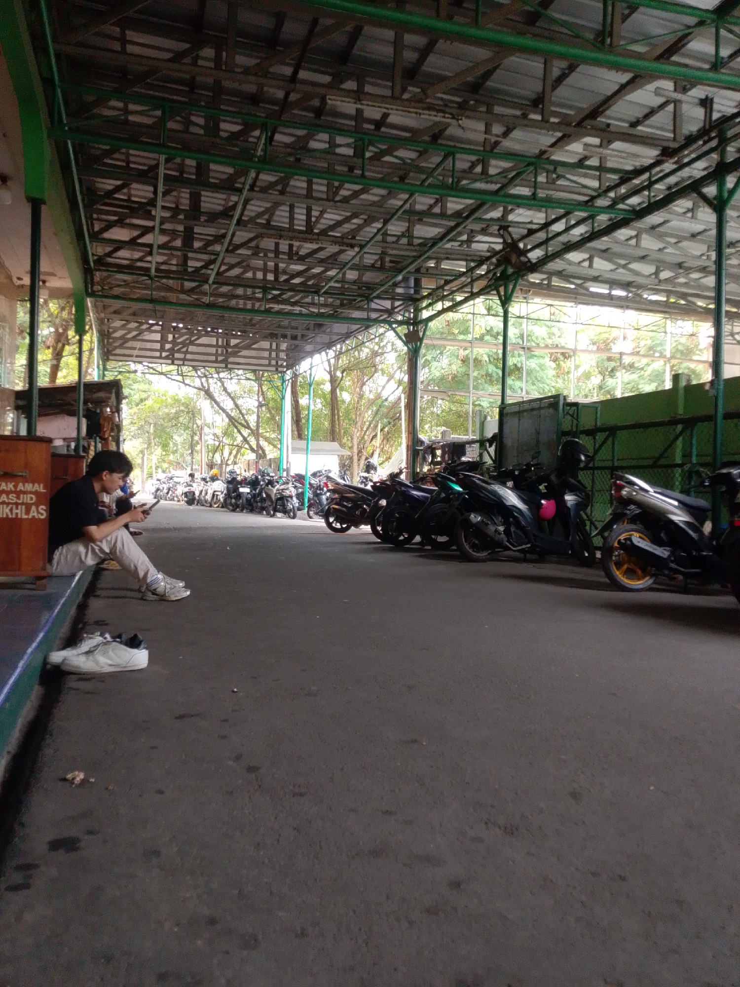 The image depicts an outdoor scene in Central Jakarta, Indonesia, featuring a covered parking area for motorcycles. A young person is seated on a ledge, looking at their phone. Several motorcycles are parked in rows under a metal canopy, suggesting a public space such as a mosque, office building, or market. The ground is paved, and trees are visible in the background, indicating an urban environment. A small wooden structure with text is partially visible to the left of the seated person.