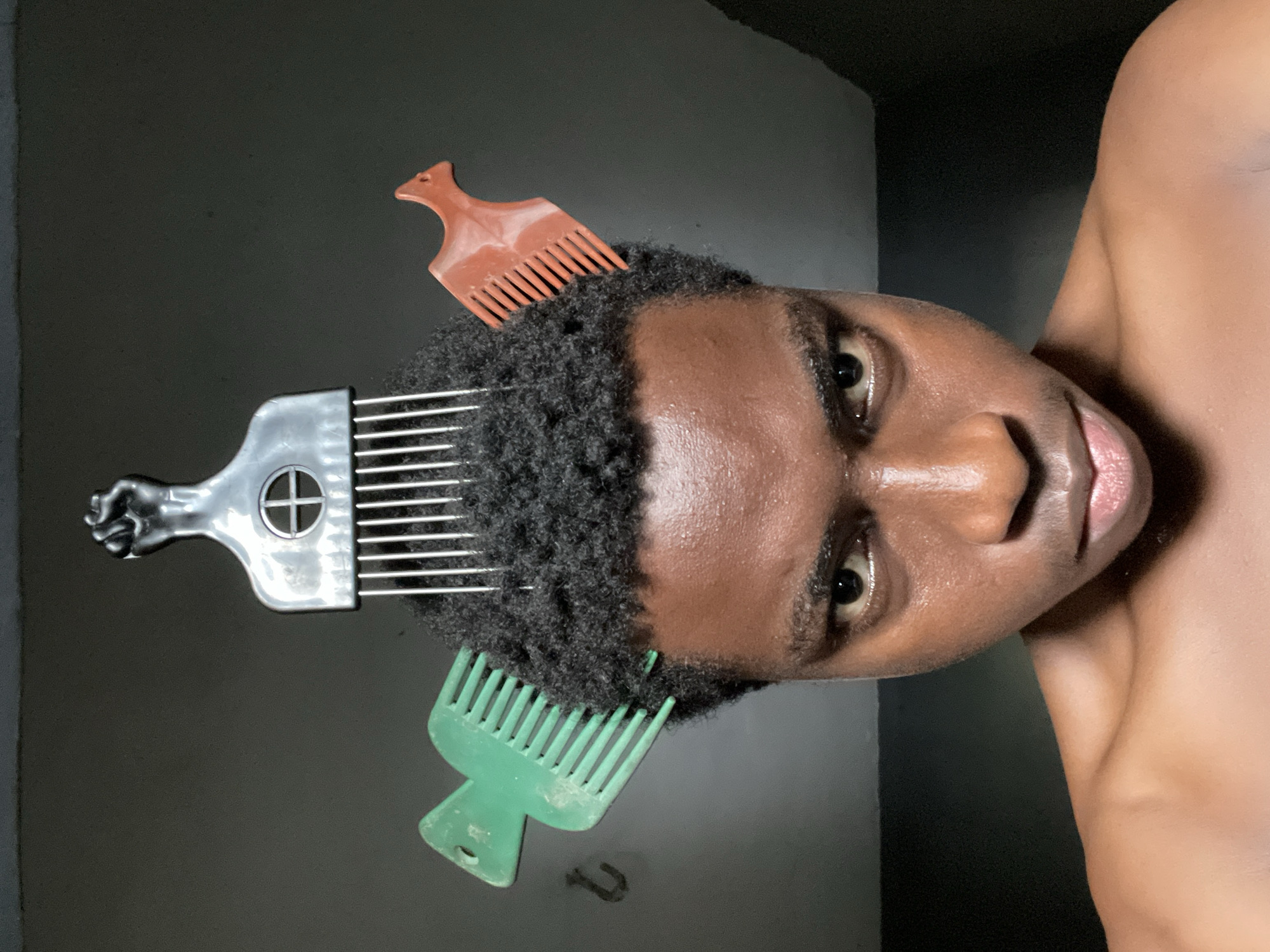 A close-up, tilted portrait shows a dark-skinned person with short, curly black hair styled with three hair picks. A large, silver-colored pick with a raised fist handle is near the top of the head, a smaller brown pick is near the upper right of the head, and a green pick is near the lower-left of the head. The background appears to be a dark gray or black surface. Part of the person's shoulder is visible on the right side of the frame. The lighting suggests a somewhat dim or artificial light source. There is no clear indication of Nkpor, Nigeria in the image.