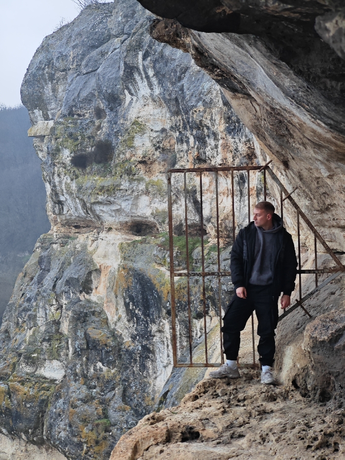 A single adult male stands on a rocky ledge at the entrance to a natural cave or rock-cut structure on the Shumen Plateau, near Shumen, Bulgaria. The male, with short light hair, is wearing a dark bomber jacket over a grey hooded sweatshirt, black cargo pants, and light-colored athletic shoes. He is positioned to the right of a rusted, vertically barred metal gate that partially obstructs the opening of the rock formation. The male is looking intently towards the viewer's left, with his body angled slightly away from the camera. To his left and behind him, a massive, stratified limestone or sandstone cliff face rises steeply, displaying varying shades of grey, beige, and traces of moss. An overhang of the cliff forms a dark ceiling directly above the subject and the gate. Distant, muted green vegetation is visible through a hazy atmosphere beyond the main cliff. The scene is illuminated by diffuse, overcast light.