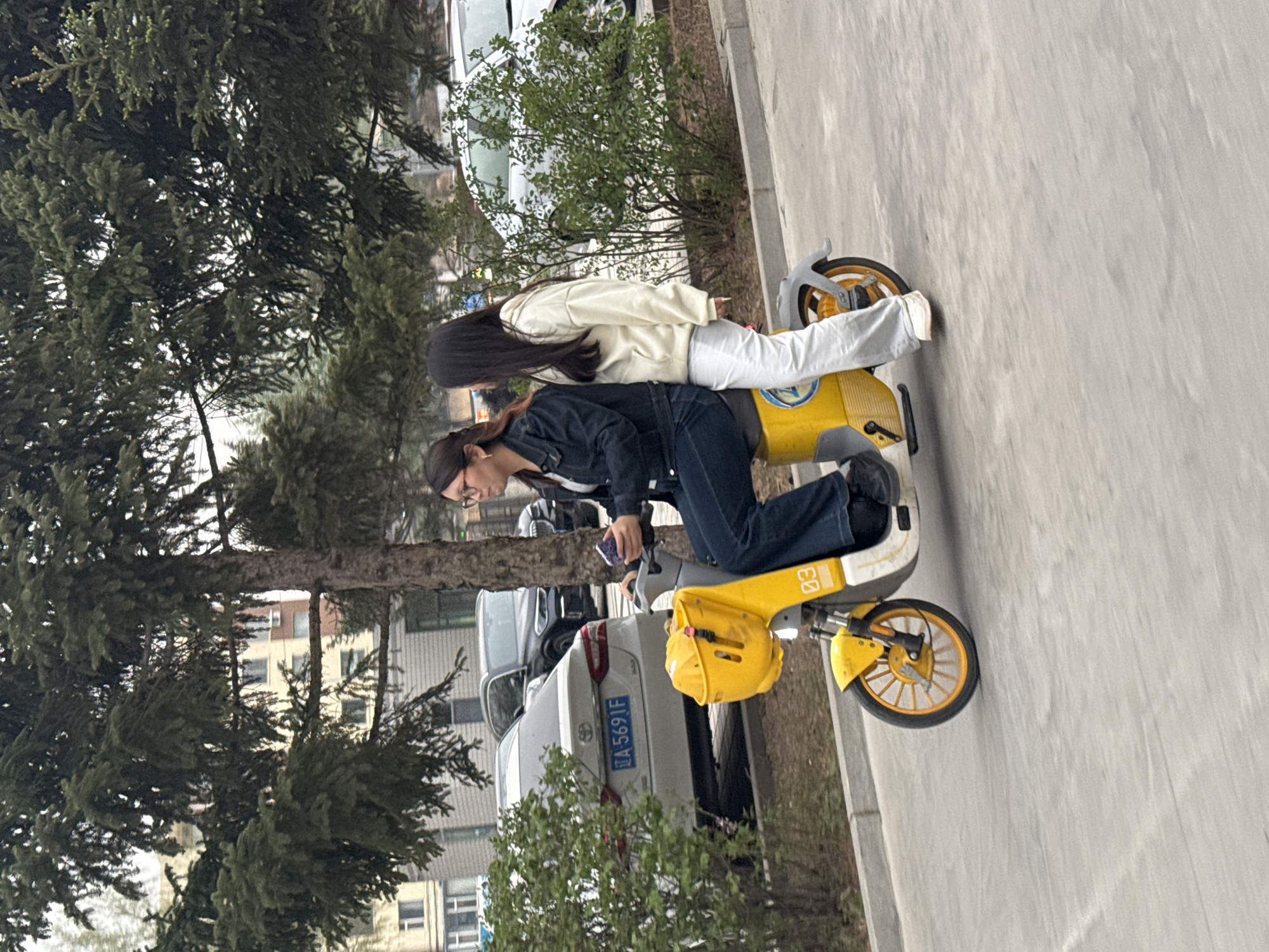 Two young women are depicted on a shared yellow scooter on a paved area, with trees and urban buildings in the background. The image appears to be taken during daylight hours on an overcast day.
One woman, wearing a dark denim jacket and dark jeans, is seated on the scooter, leaning forward slightly as she looks down at a phone held in her left hand. Her hair is dark and pulled back. The second woman stands behind her on the scooter's platform, dressed in a light-colored (possibly cream or off-white) hooded sweatshirt and light-colored pants, with long dark hair flowing down her back. Both appear to be focused on the activity with the scooter.
The scooter itself is bright yellow with gray accents and has "03" visible on its side, indicating it is likely part of a shared mobility service. It features a small basket at the front. The women are positioned on what appears to be a sidewalk or pathway, with a darker paved road adjacent to it.
In the background, lush green trees, possibly evergreens, line the path. Beyond the trees, several parked cars are visible, including a white sedan with a visible license plate that reads "A 5691F". Further in the background, multi-story buildings of varying architectural styles can be seen. The scene suggests a relaxed urban or suburban environment, consistent with Zhongtun, China.
