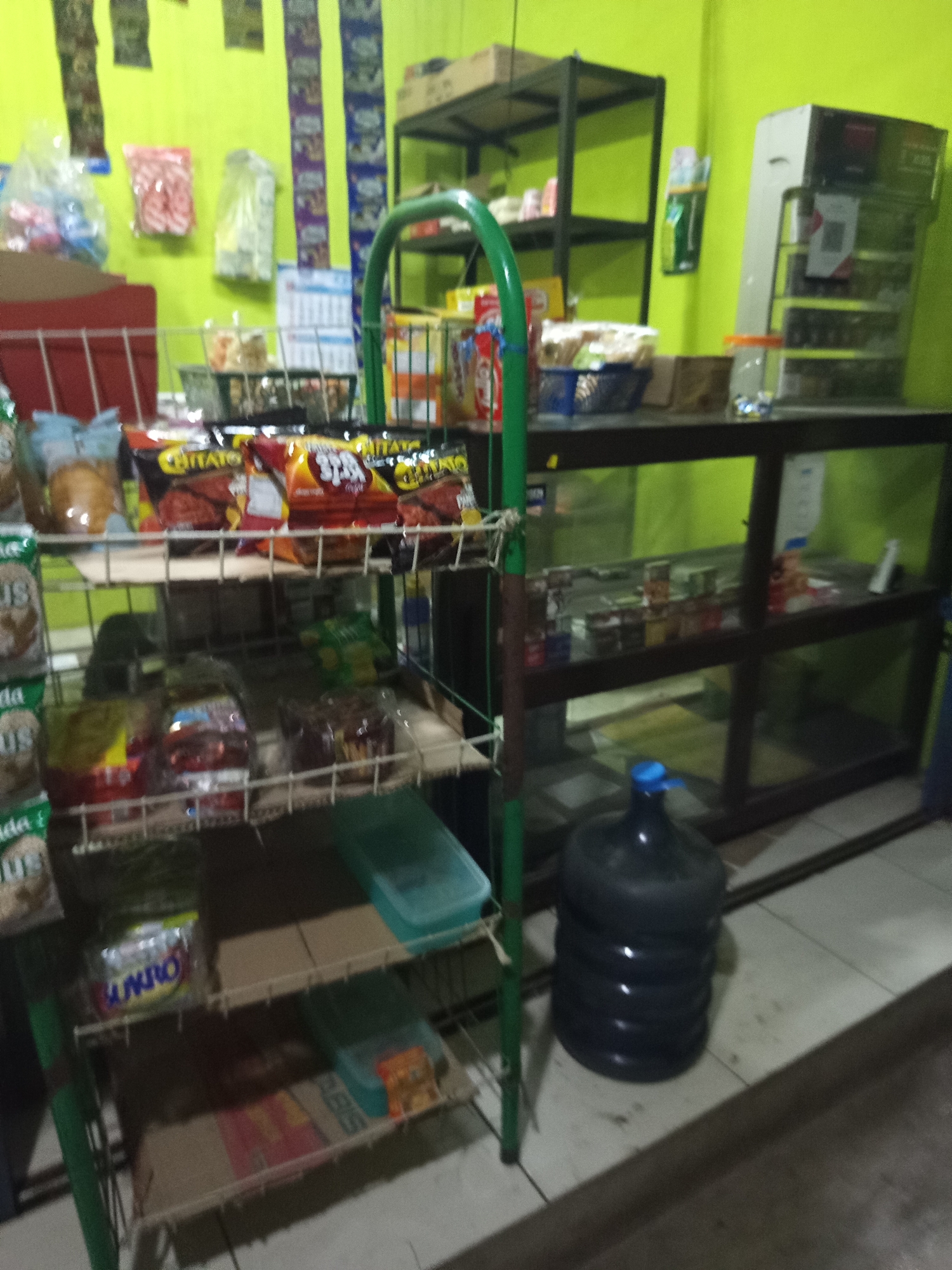 The image depicts the interior of a small retail store, possibly a warung or convenience store, located in Jayamukti, Indonesia. The walls are painted a vibrant lime green, and the floor appears to be tiled.
In the foreground, a metal wire rack displays various products. The top shelf holds bagged snacks, including brightly colored packets with Indonesian writing. Some of these appear to be "Chitato" brand chips. Below this, another shelf contains more bagged items, some of which are translucent, revealing their contents. The lower shelves are mostly obscured by shadows, but one clearly shows a package with the word "SUNGO" in blue lettering. To the right of the shelving unit is a large, dark blue water cooler bottle with a blue cap, sitting on the floor.
In the background, to the left, is a red plastic bin or display. Further back, a black metal shelving unit is visible, holding various items, including what appears to be more bagged products. To the right of this unit, a glass-fronted display cabinet is filled with rows of small items, likely packaged goods or food products. A white dispenser is mounted on the wall behind the cabinet. The overall impression is that of a well-stocked, albeit somewhat cluttered, small retail space.