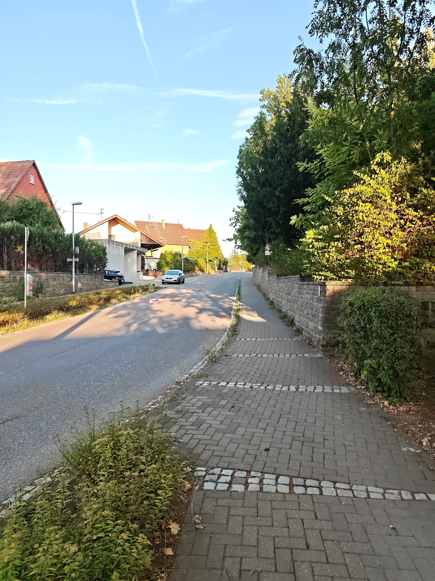The image shows a street scene in Nordheim, Germany. The foreground features a paved sidewalk with a pattern of grey interlocking pavers and a decorative border of smaller cobblestones. To the left of the sidewalk is a narrow strip of green foliage. The sidewalk runs alongside a grey asphalt road.
On the right side of the road, a low stone wall runs parallel to the sidewalk, topped with dense green bushes and trees. Further down the road, on the left side, are several houses with red-tiled roofs and a modern white building. Two cars are visible on the road: a dark-colored car parked near the white building and a silver car driving away from the camera. The sky above is clear blue with a contrail from an aircraft and some wispy white clouds. The lighting suggests it is late afternoon or early evening. A speed limit sign with a "30" is visible on the right side of the road, near the stone wall.