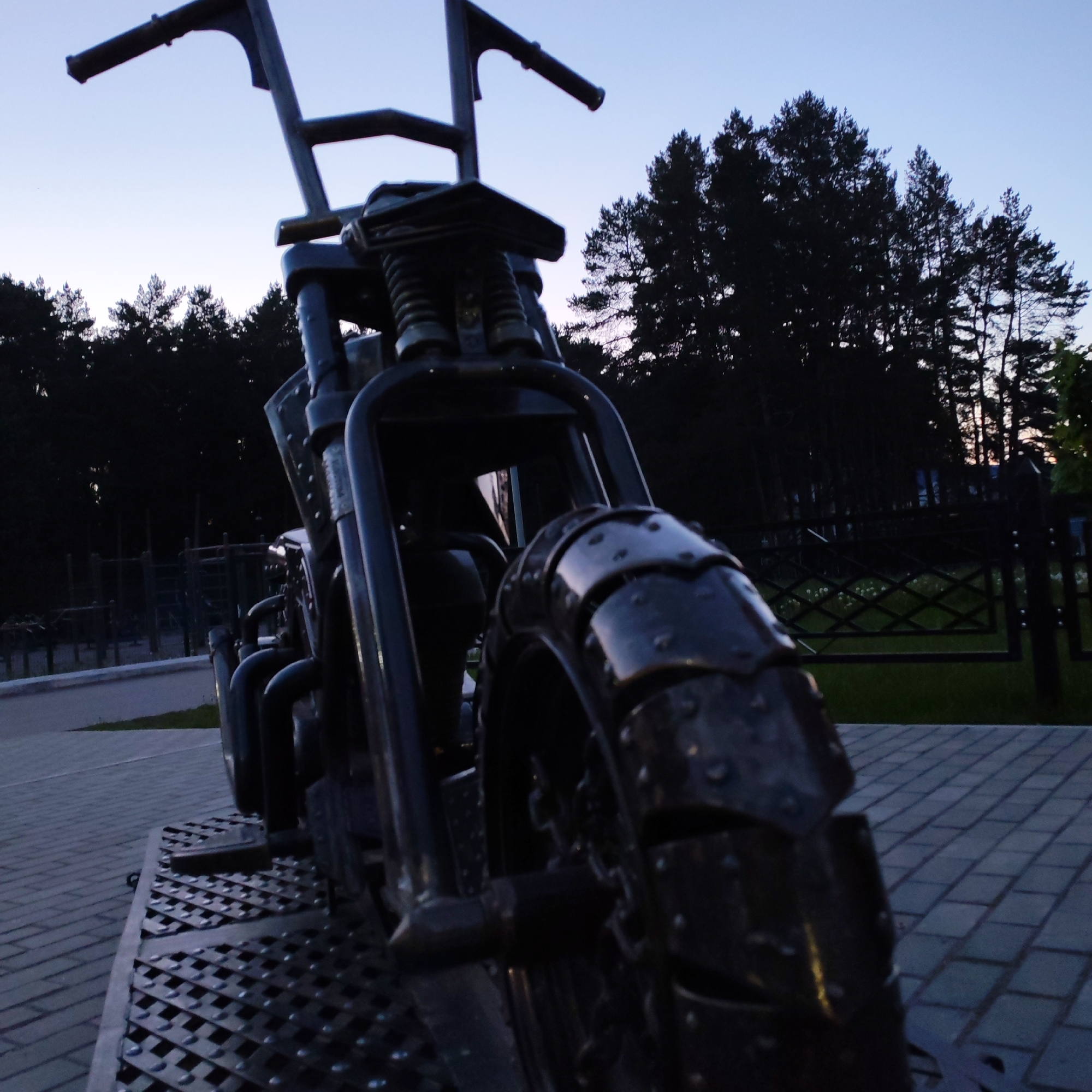 A low-angle, full shot shows a metal sculpture of a motorcycle in the city of Chaikovsky, Russia. The sculpture features a large front tire with studded metal plates. The handlebars are high-mounted, and the overall design is rugged. The motorcycle is on a metal grate platform, which sits on a brick-paved surface. A dark row of trees forms the backdrop against a twilight sky. A metal fence is visible in the background. The overall tone is subdued, due to the low light and dark colors. No people are visible.
