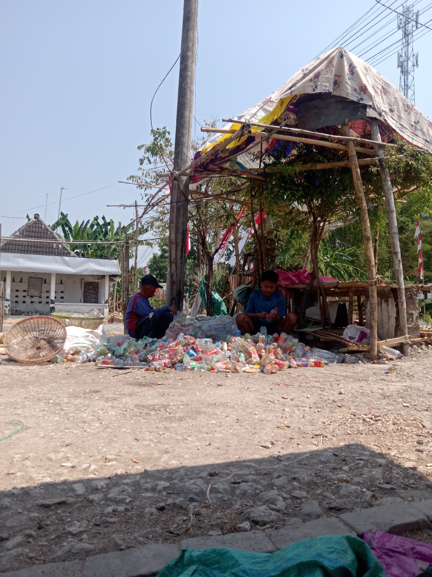 In the city of Stiaji, Indonesia, two individuals are sorting through a large collection of discarded plastic bottles. They are situated in an outdoor area with a dirt and gravel ground. A makeshift shelter, constructed from bamboo poles and covered with various fabrics, provides shade. One person, wearing a blue shirt, is seated under the shelter, holding a plastic bottle and appearing to examine it. The other individual, dressed in a blue shirt and red vest, is crouched outside the shelter, surrounded by a considerable amount of plastic bottles. A woven basket and a pile of white sacks are also visible nearby. In the background, a modest building with a tiled roof and some vegetation, including banana trees, can be seen. The scene is illuminated by natural daylight under a clear sky.
