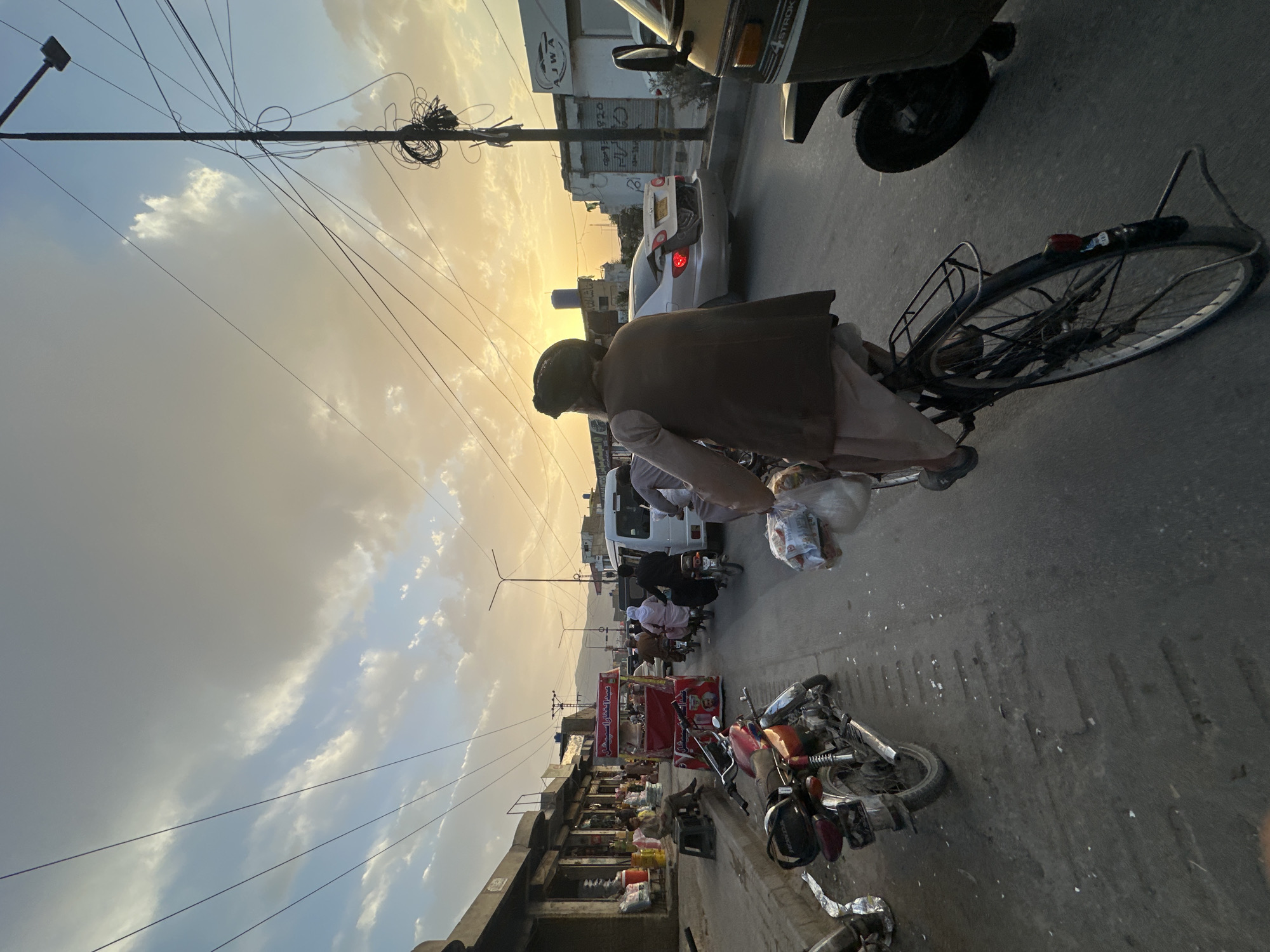 The image shows a street scene in Quetta City Tehsil, Pakistan, at sunset. The sky is a mix of blue and cloudy with a yellow glow. Numerous overhead electrical wires extend across the frame. The street is lined with shops and vendor stalls. People are on foot and on motorbikes and bicycles. A man wearing a traditional pakol hat and vest is riding a bicycle with a bag of goods hanging from his handlebars. Several parked motorbikes are visible along the side of the road. An auto-rickshaw and a white car are in the background. The scene is typical of a busy marketplace.