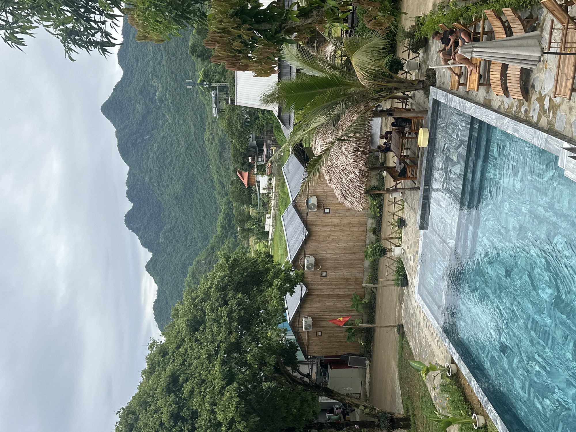 The image depicts an outdoor scene with a swimming pool in the foreground, surrounded by lounge chairs and a thatched-roof seating area. Beyond the pool, there are several small bamboo-built structures, some with air conditioning units. A Vietnamese flag is visible on a flagpole. In the background, lush green mountains rise under a cloudy sky, with some scattered buildings and trees visible at their base. Three individuals are seated in lounge chairs by the pool, one of whom appears to be on their phone. Another group of people is seated at a table under the thatched roof. The location is identified as Xóm Khán, Vietnam.