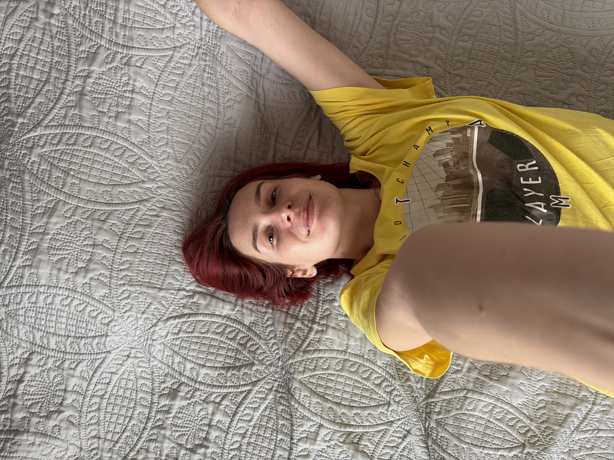 A young woman is depicted in a high-angle selfie, lying comfortably on a light-colored, intricately quilted surface, possibly a bed or sofa, in Chaikovsky, Russia. She has shoulder-length, reddish-brown hair and a calm expression with a subtle smile. Her left arm is extended upwards and partially out of frame, while her right arm, holding the camera, stretches towards the viewer in the foreground.
She wears a bright yellow t-shirt featuring a large circular graphic. Visible text on the shirt includes "C H A M P" arcing on the left side of the circle, and "LAYER M" printed vertically on the right. The graphic itself appears to be a muted-toned illustration of a cityscape or buildings.
The quilted surface beneath her is light grey or off-white, decorated with repeating patterns of stitched floral or leaf-like motifs and geometric shapes. The scene is well-lit by bright, soft natural light, indicating it is daytime. The overall mood conveyed is one of relaxation and contentment.